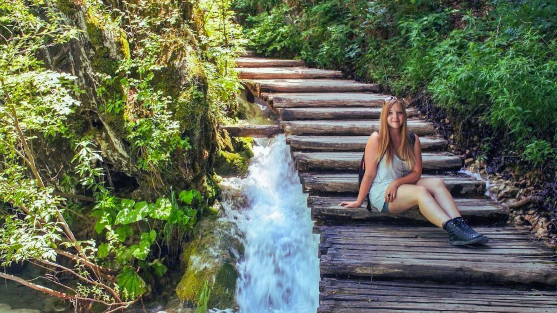 A woman smiles while sitting on a wooden path beside a cascading stream in a lush forest, conveying a sense of tranquility and nature's beauty.