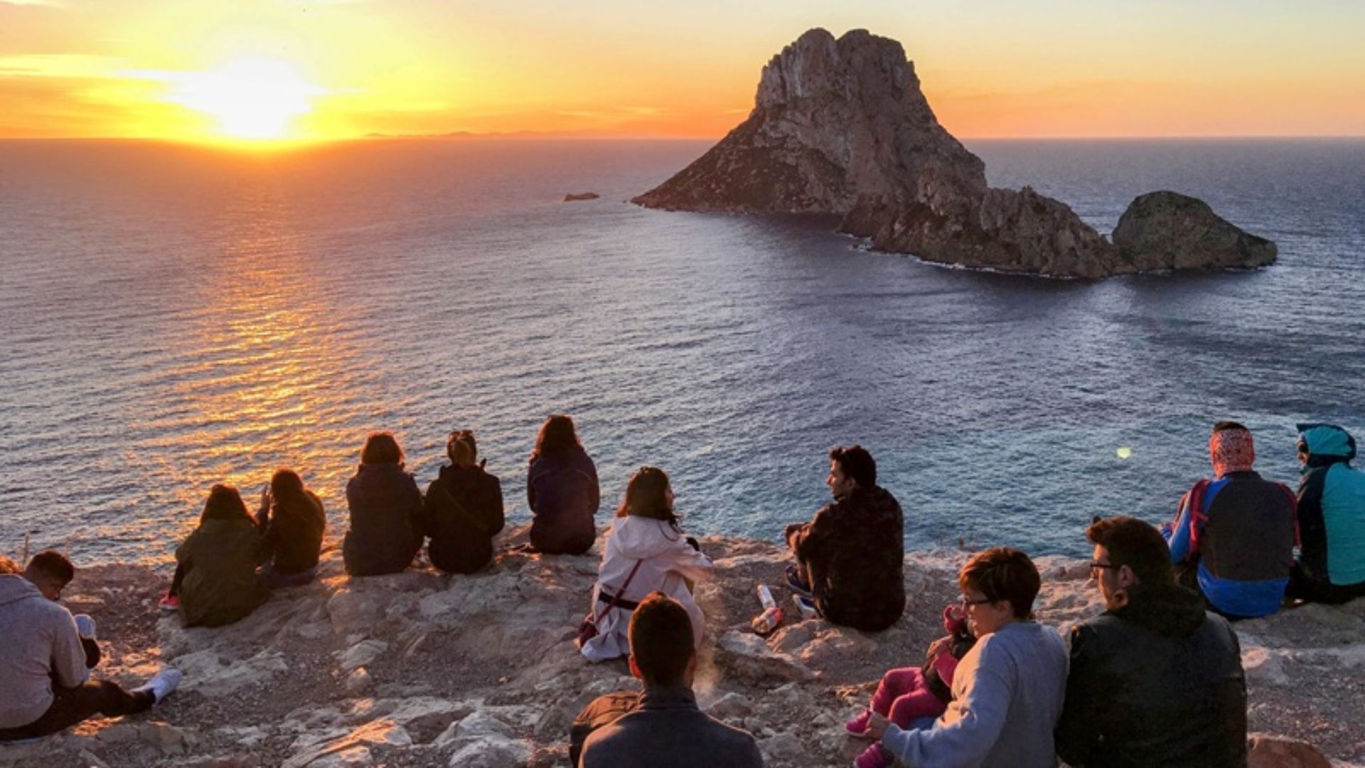 A group of people sit on a rocky cliff, watching a serene ocean sunset. An island silhouette is visible in the distance. The scene is peaceful and contemplative.