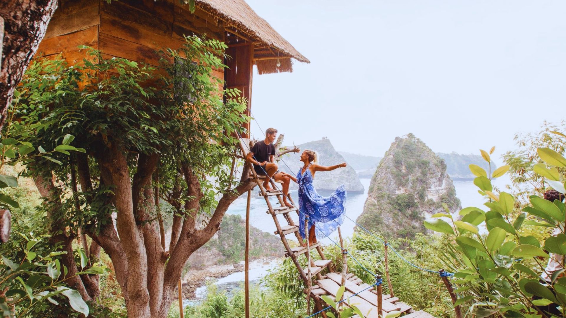 A couple happily enjoys a scenic view from a treehouse. The woman in a blue dress stands on a ladder, surrounded by lush greenery and cliffs over a bright sea.