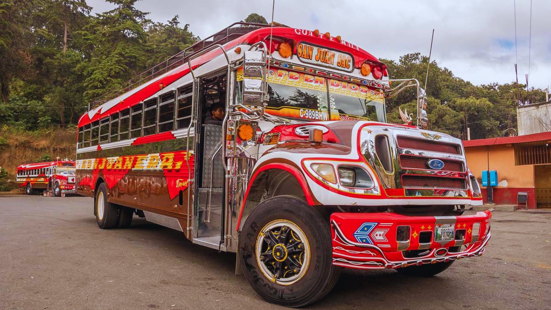 A vibrant, custom-painted bus features bold red and white patterns with chrome accents, parked on a street with trees and a blue sky backdrop.