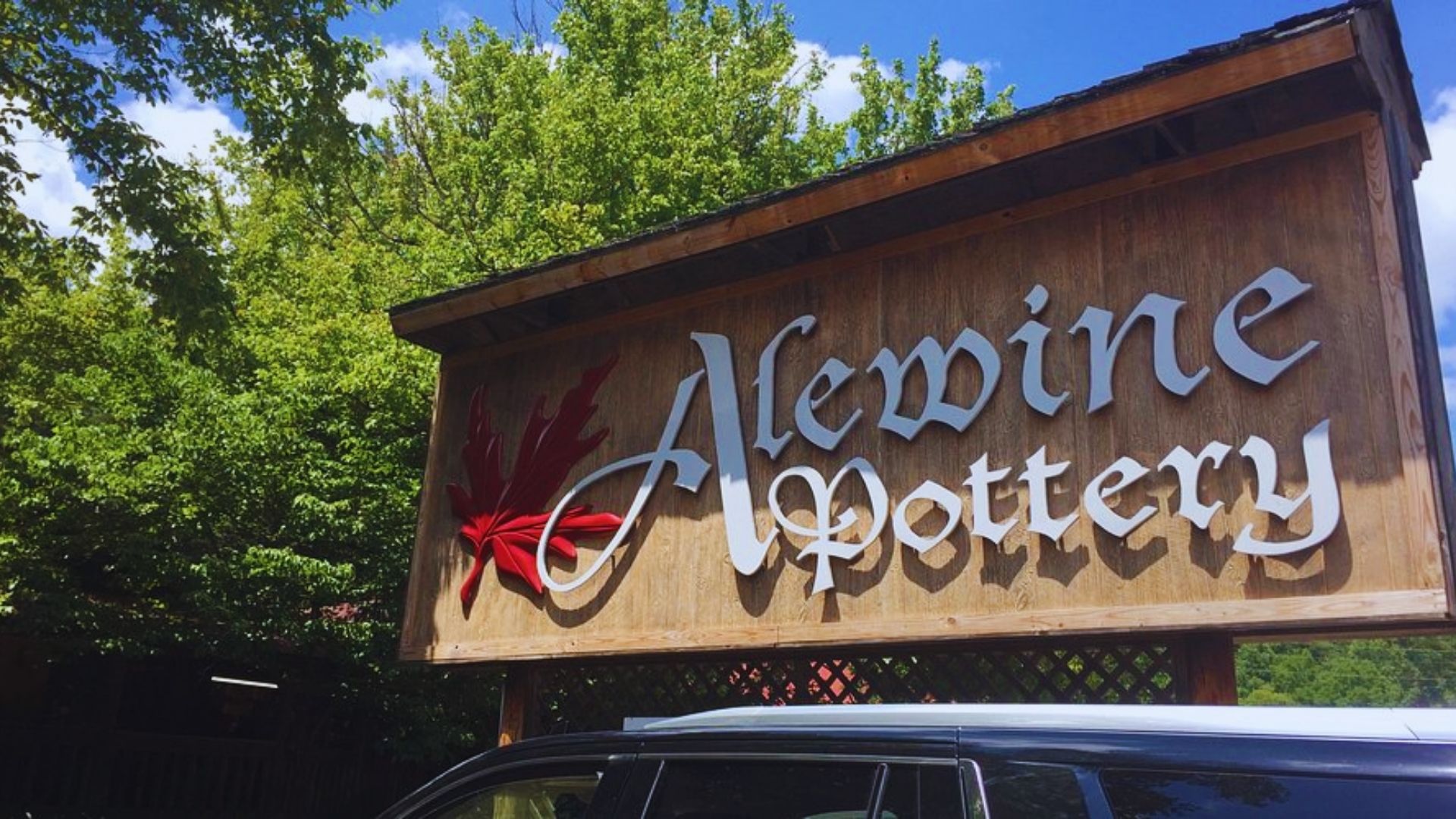 Wooden sign reading "Alewine Pottery" with decorative red leaf, set against green trees and a blue sky. Car roof visible below the sign, conveying a rustic, artistic vibe.