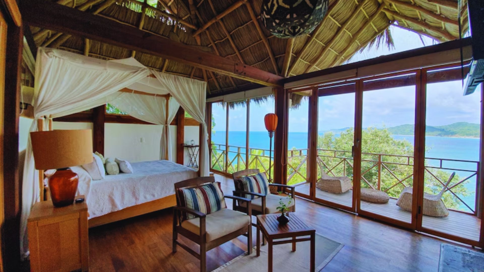 Tropical bedroom with wooden decor and a canopy bed, overlooking a blue ocean. Large windows open to a scenic balcony, creating a serene ambiance.