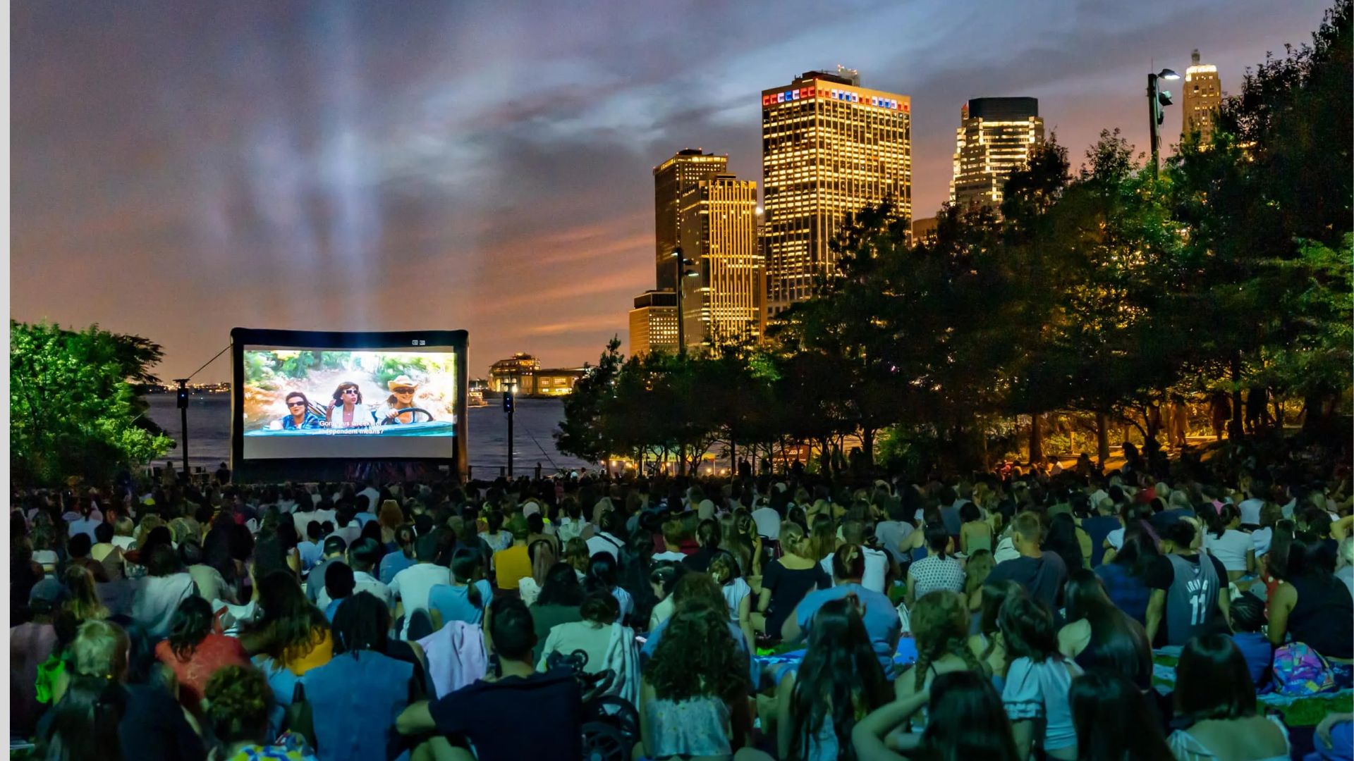 A large crowd gathered outdoors, intently watching a movie projected on a big screen at night.