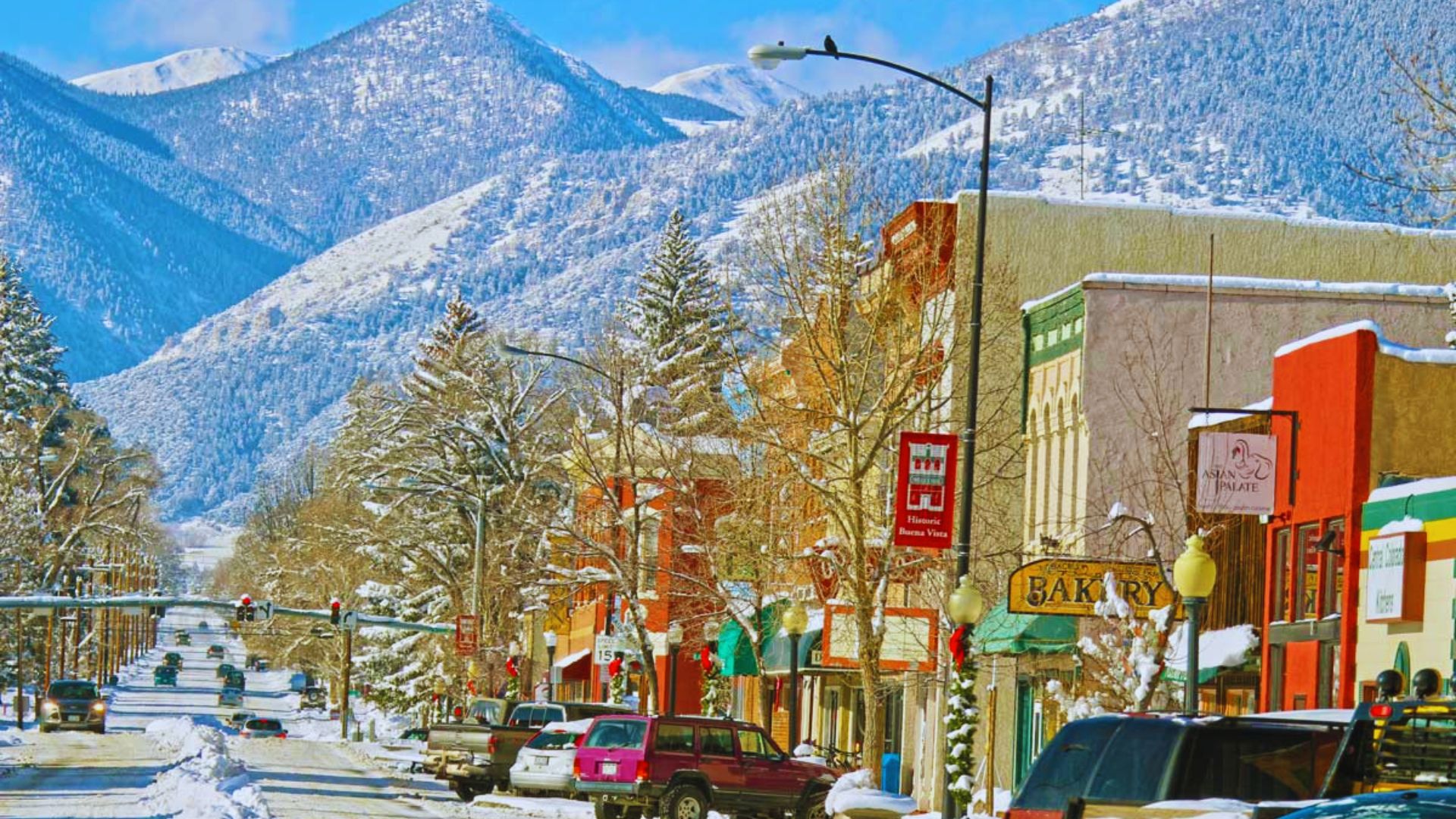 Snow-covered street with parked cars, leading to a majestic mountain in the background.