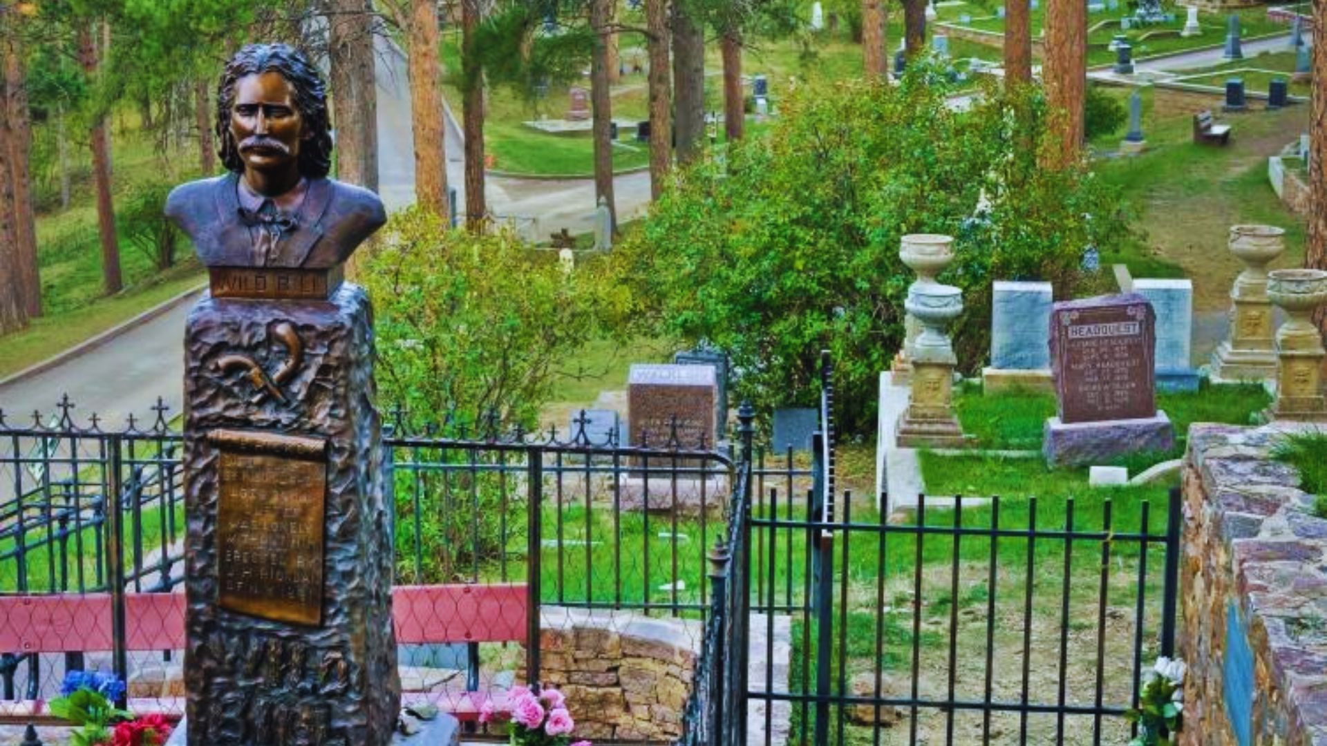A man’s statue in a cemetery, accompanied by flowers and bordered by a fence.