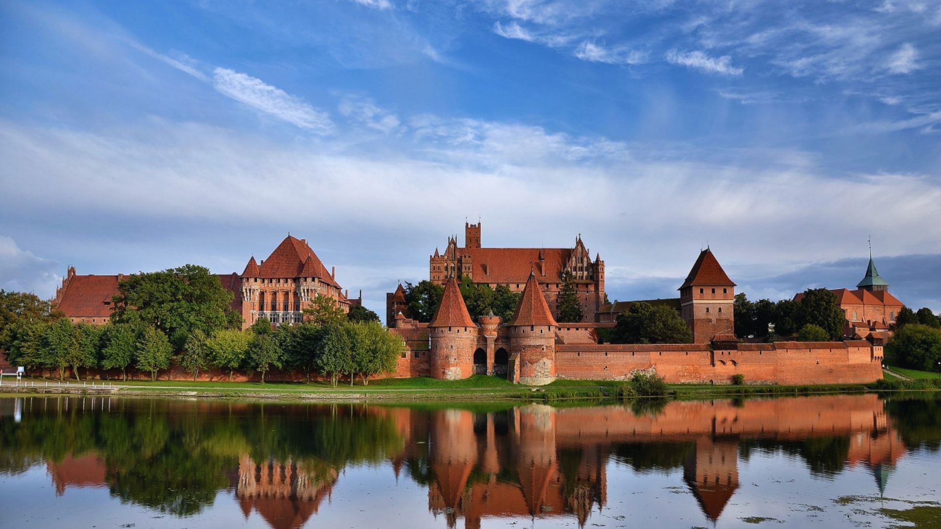 A grand medieval castle with red brick walls and conical roofs reflects on a calm lake. Surrounded by lush trees, the scene conveys historical majesty.