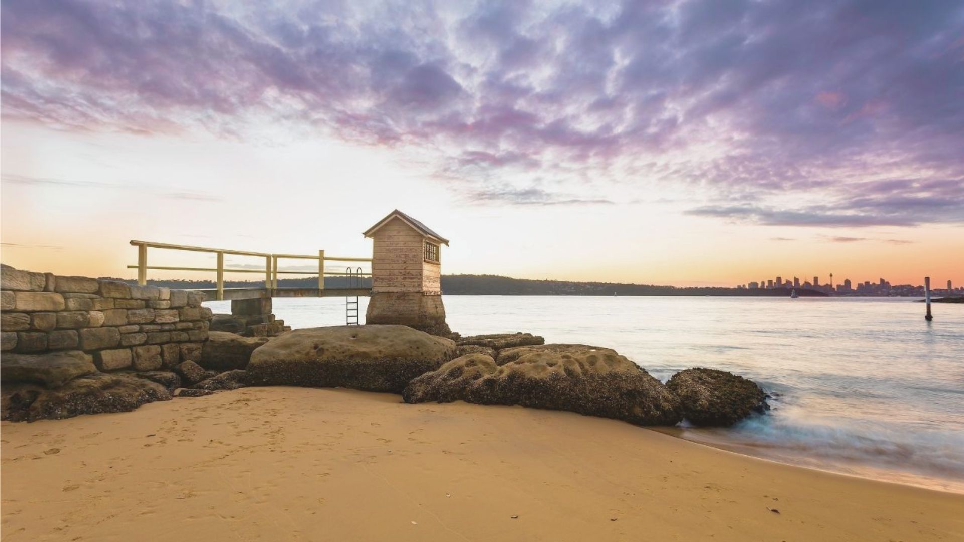 A small wooden shed stands on a rocky outcrop at sunset, bordered by calm waters and a sandy beach. The sky is vibrant with pink and purple hues.