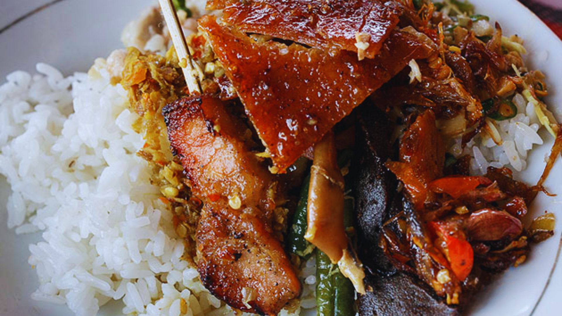 A plate of white rice topped with vibrant Balinese mixed vegetables and crispy, flavorful roast pork. The dish looks savory and indulgent.