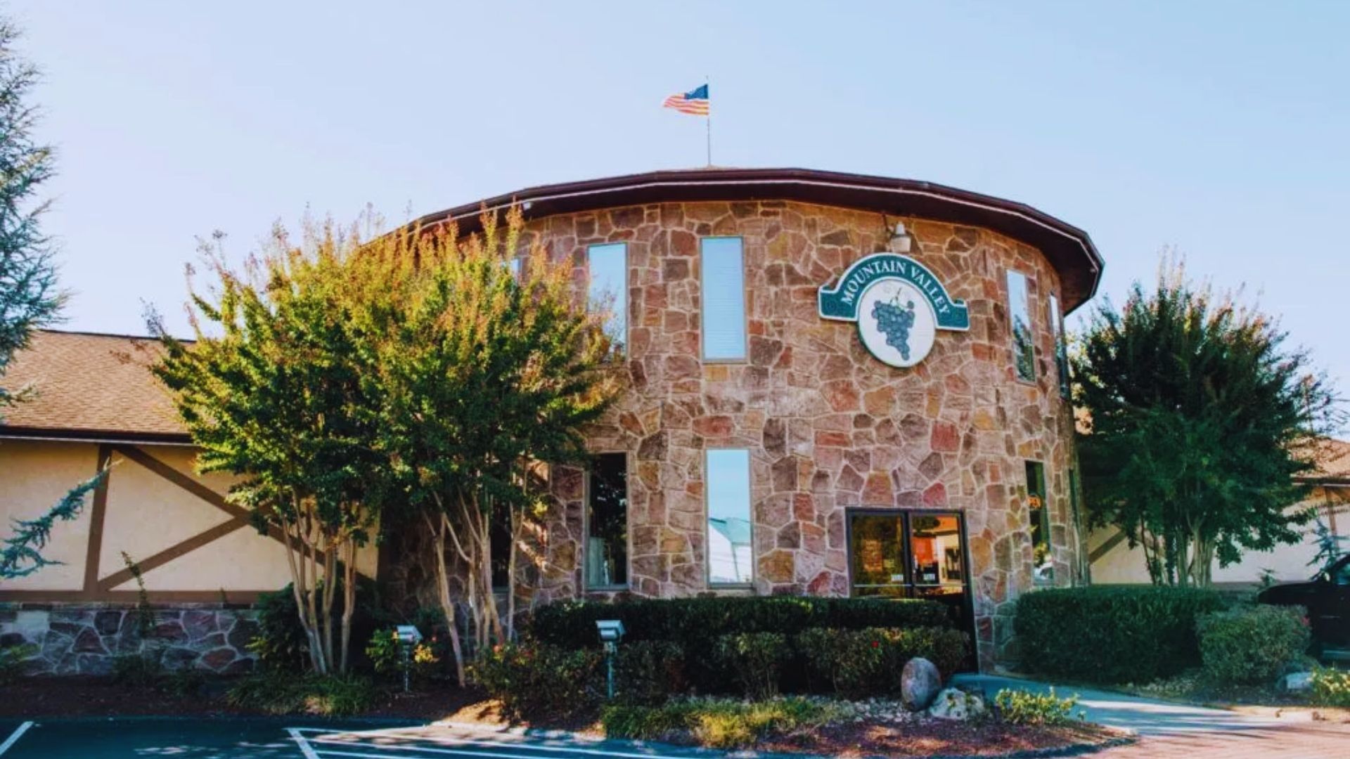 A stone building with tall windows, an American flag on top, and a sign reading "Mountain Valley." Surrounded by trees and bushes, under a clear sky.