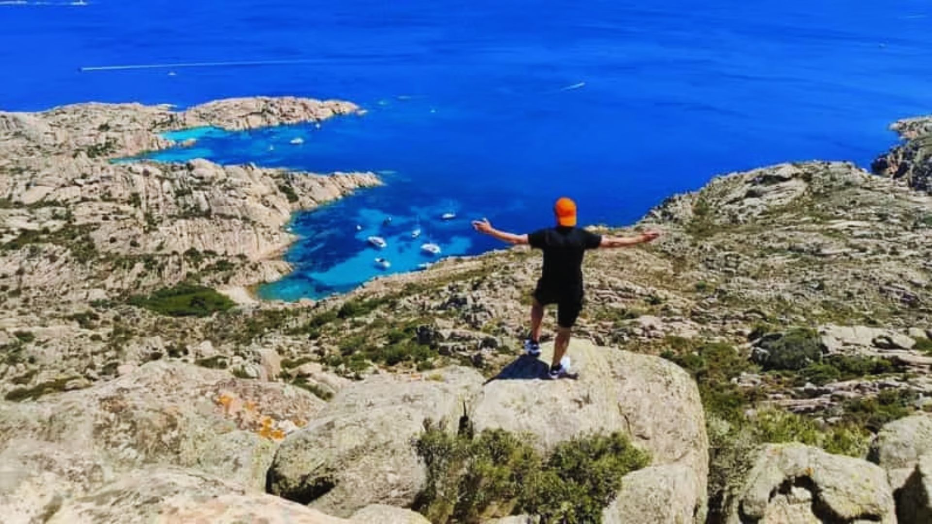 A person in a black outfit and orange cap stands on a rocky cliff overlooking a stunning blue coastline with scattered sailboats, arms outstretched in awe.