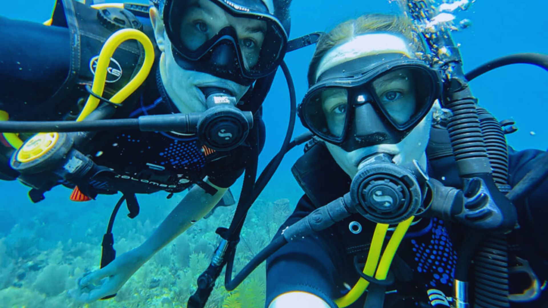 Two scuba divers smiling and taking a selfie underwater, surrounded by colorful fish and coral.