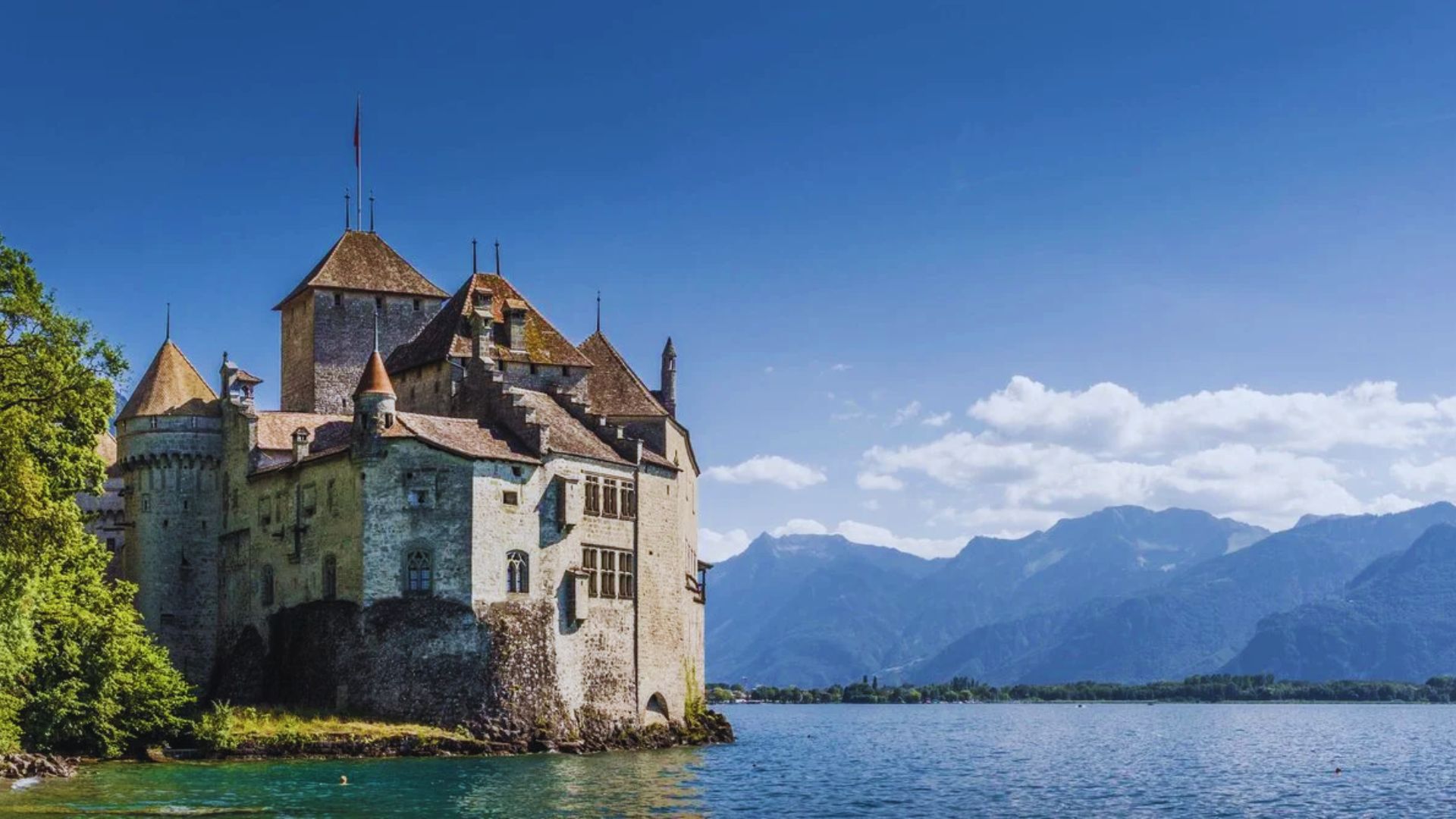 A historic stone castle with conical roofs sits by a tranquil lake, surrounded by lush greenery and distant mountains under a clear blue sky.