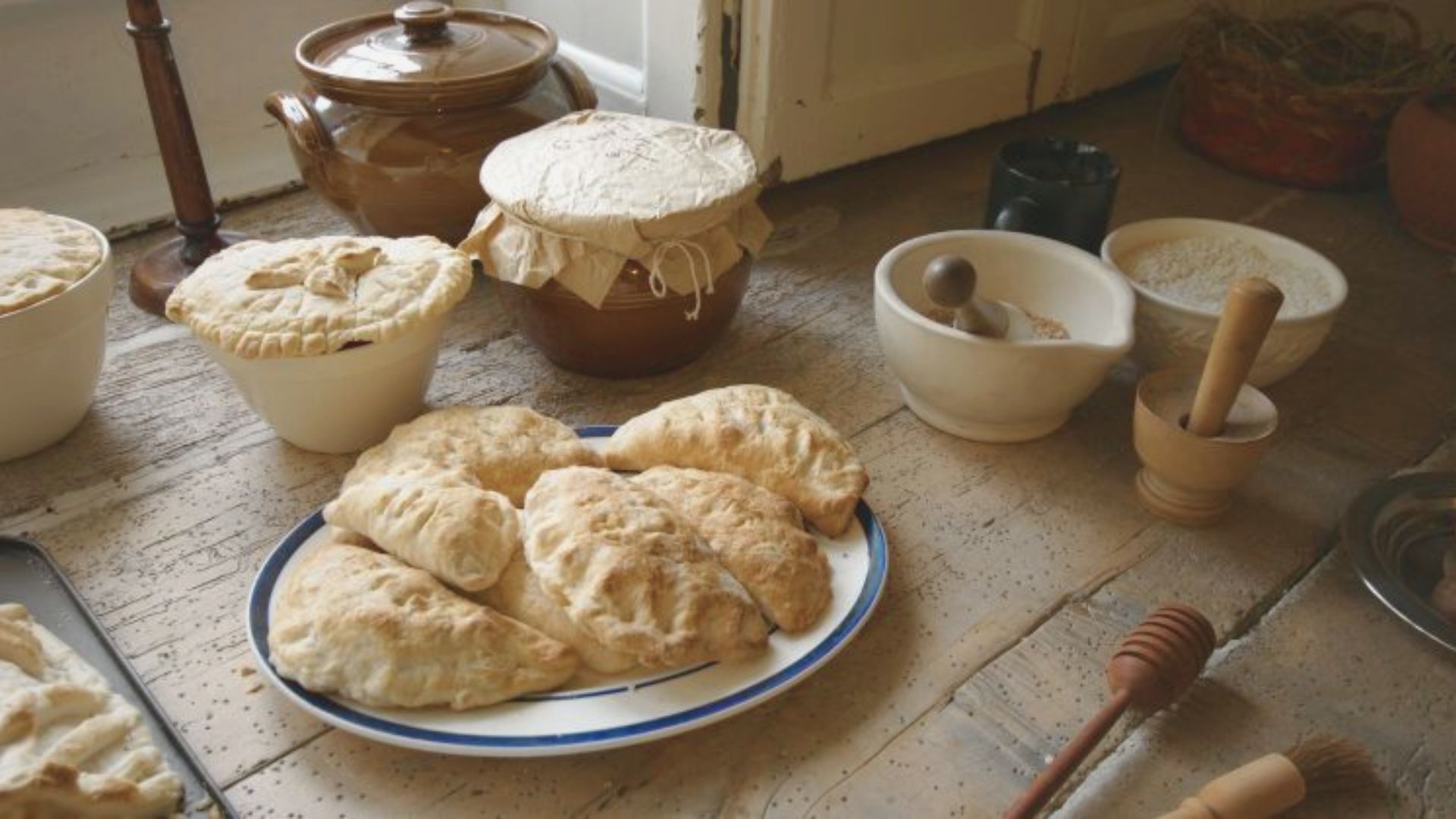 Rustic kitchen setting with freshly baked goods on a wooden table. A blue-rimmed plate holds several golden-brown pastries. Nearby are flour bowls, a honey dipper, and a lidded jar, conveying a cozy, homemade atmosphere.