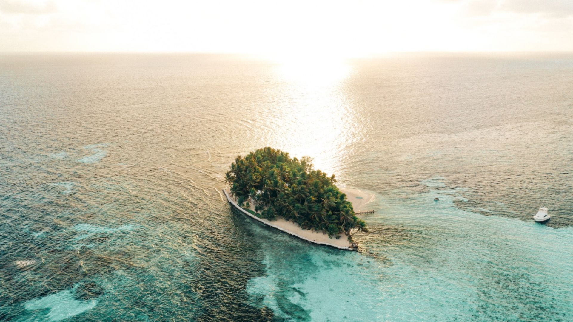 Aerial view of a small, lush tropical island surrounded by turquoise waters at sunset, evoking a serene and peaceful atmosphere. A boat floats nearby.