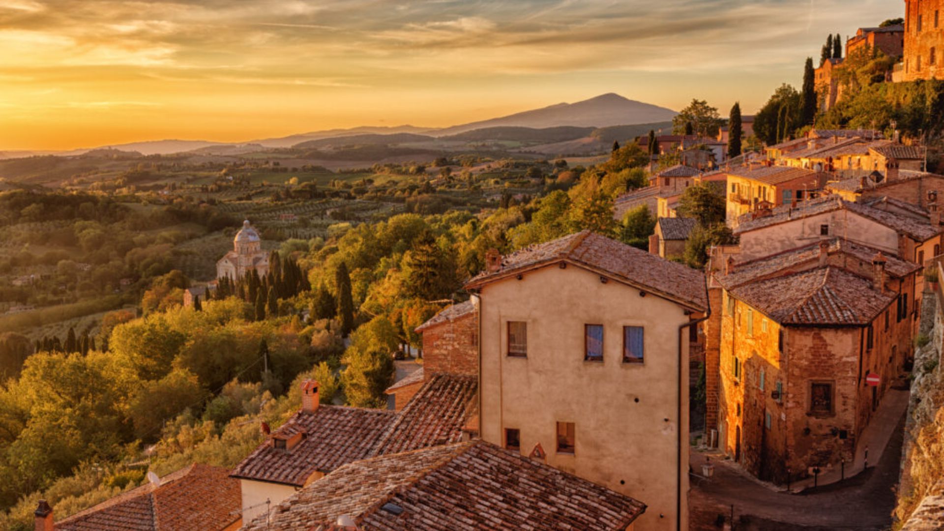 Sunset over a Tuscan village; warm golden light bathes terracotta rooftops, lush green hills, and a distant dome. Serene and picturesque landscape.