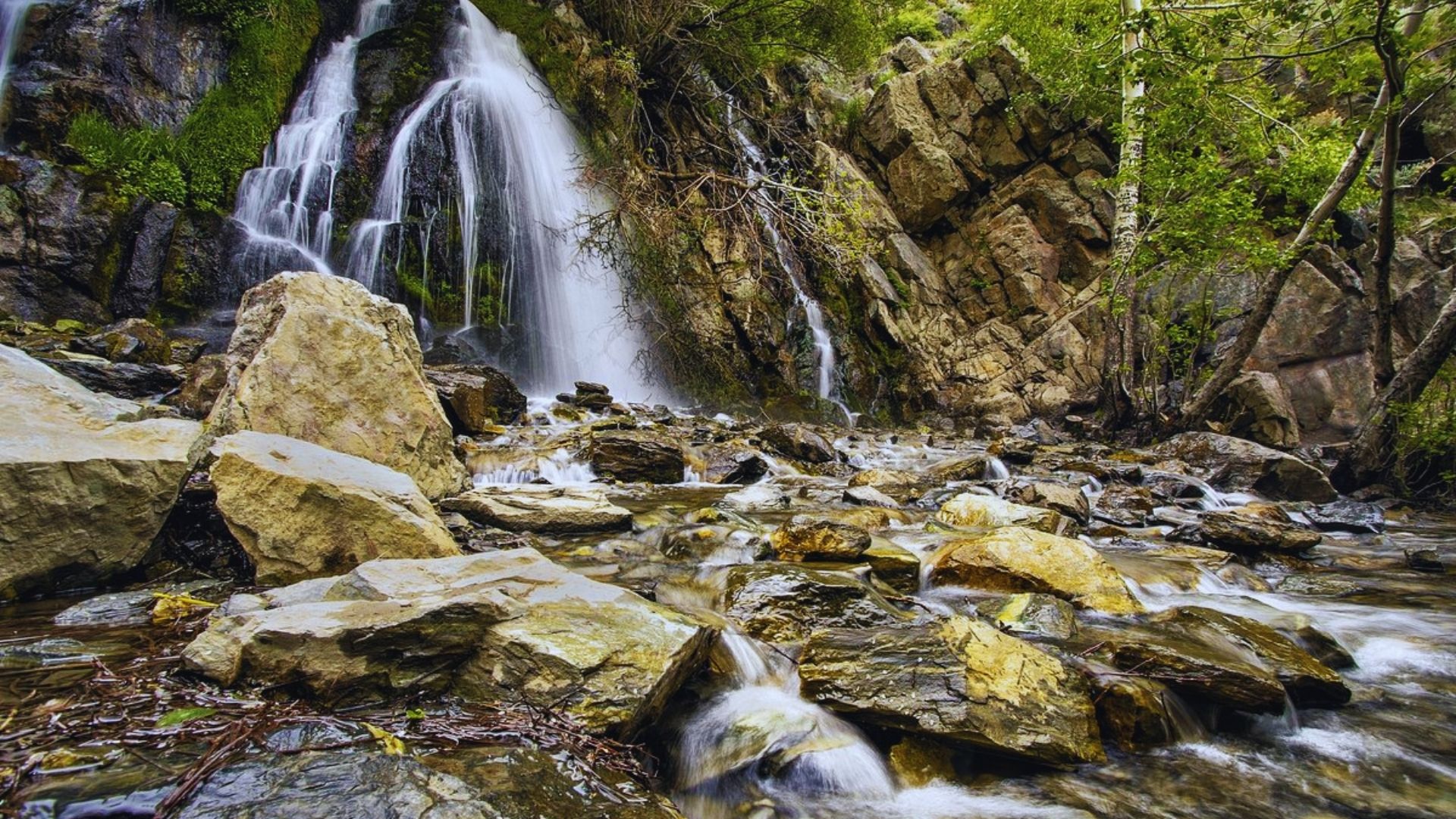 A scenic mountain waterfall cascading over rocks, surrounded by lush greenery and flowing water.