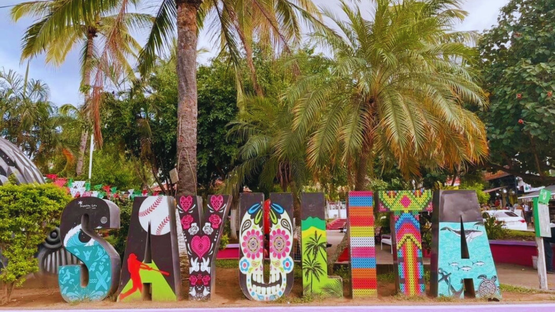 Colorful large letters spell out "SAYULITA" adorned with tropical, floral, and artistic patterns. Surrounded by palm trees, the scene is vibrant and lively.