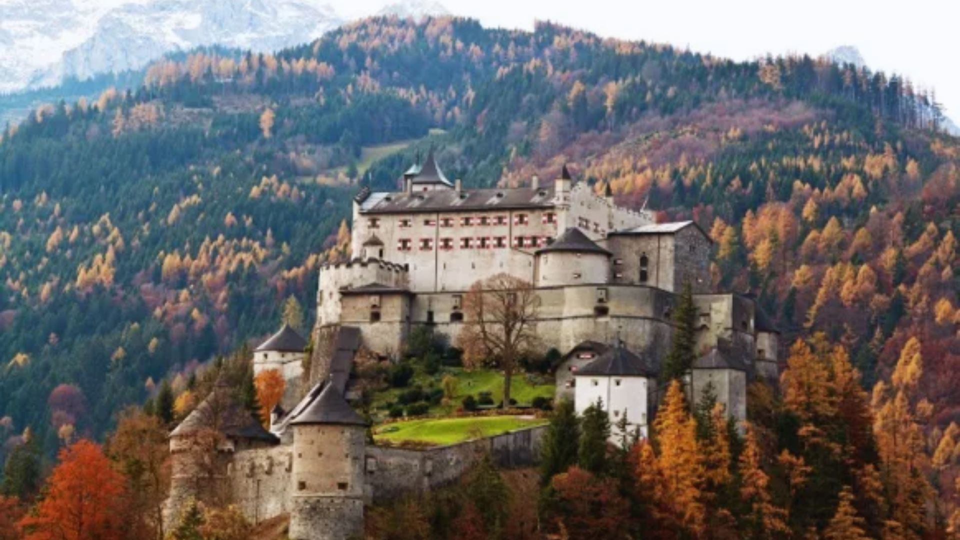 A majestic castle sits atop a hill, surrounded by autumnal trees with vibrant orange and yellow leaves, set against a backdrop of towering mountains.