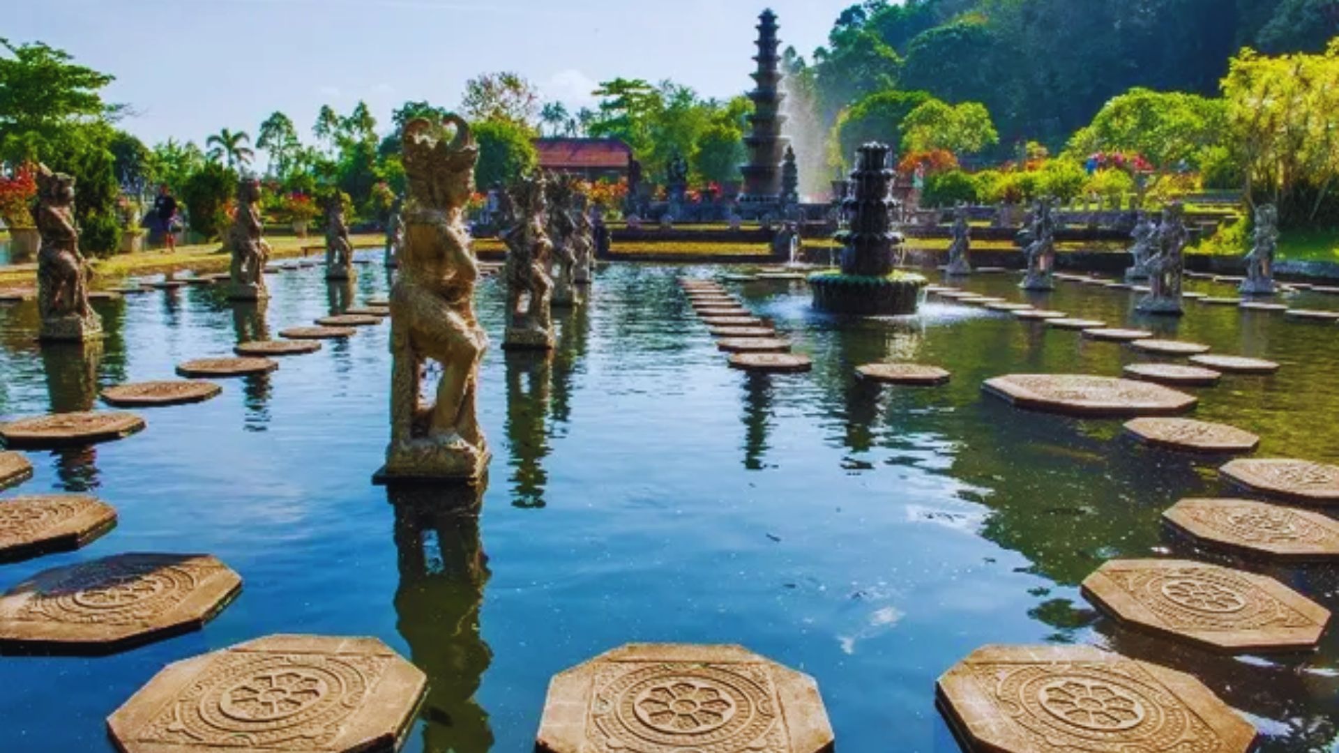 Stone statues and stepping stones adorn a serene water garden surrounded by lush greenery. In the background, a tiered fountain gracefully sprays water.