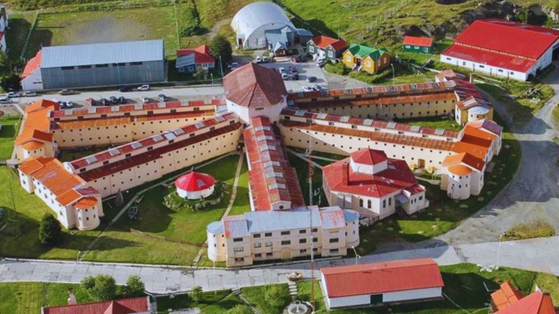 Aerial view of a historic prison complex with a star-like layout. The buildings have red roofs and beige walls, surrounded by green grass and roads. The scene conveys a sense of history and isolation.