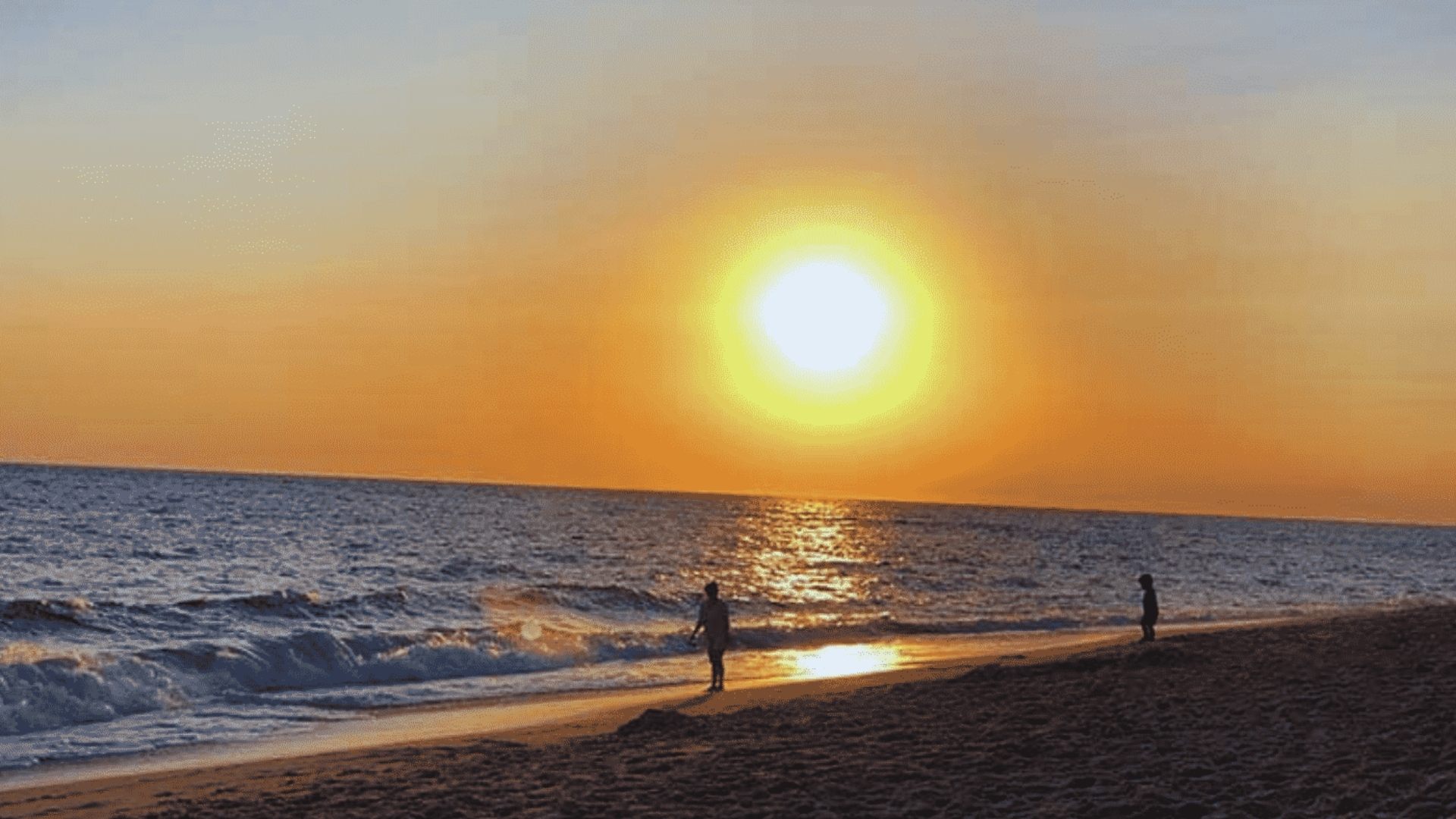 A person walks along the beach at sunset, with vibrant orange and pink hues reflecting on the water.