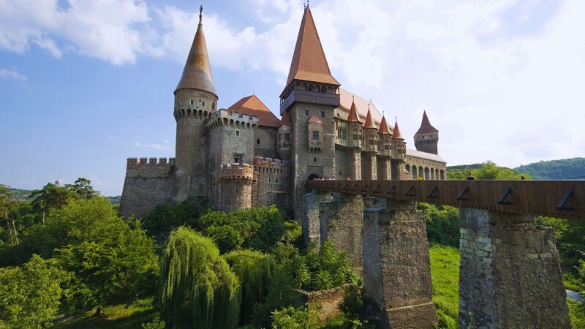 A medieval castle with tall, pointed towers and a bridge spans lush green hills. The sky is partly cloudy, creating a serene, historic ambiance.