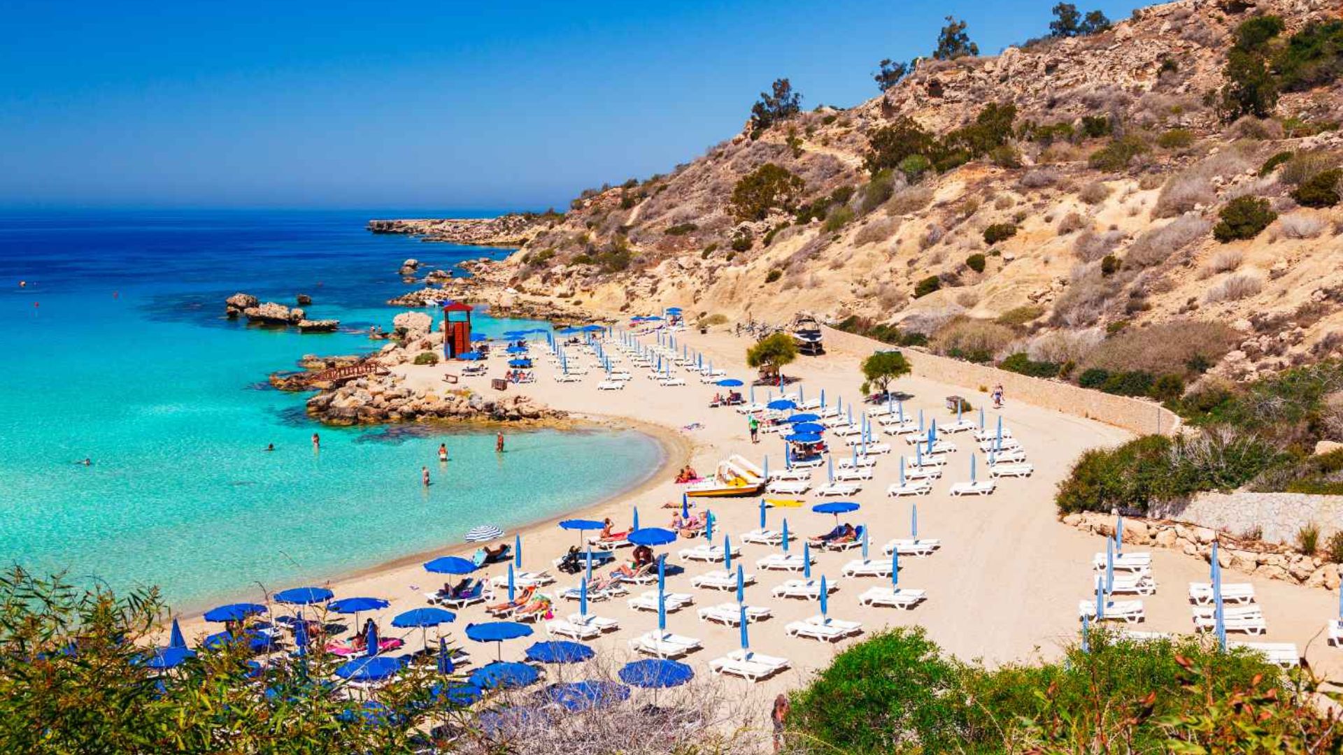 A picturesque beach scene with turquoise waters and a sandy shore lined with rows of blue umbrellas and white lounge chairs. Rocky hills meet the clear blue sky.
