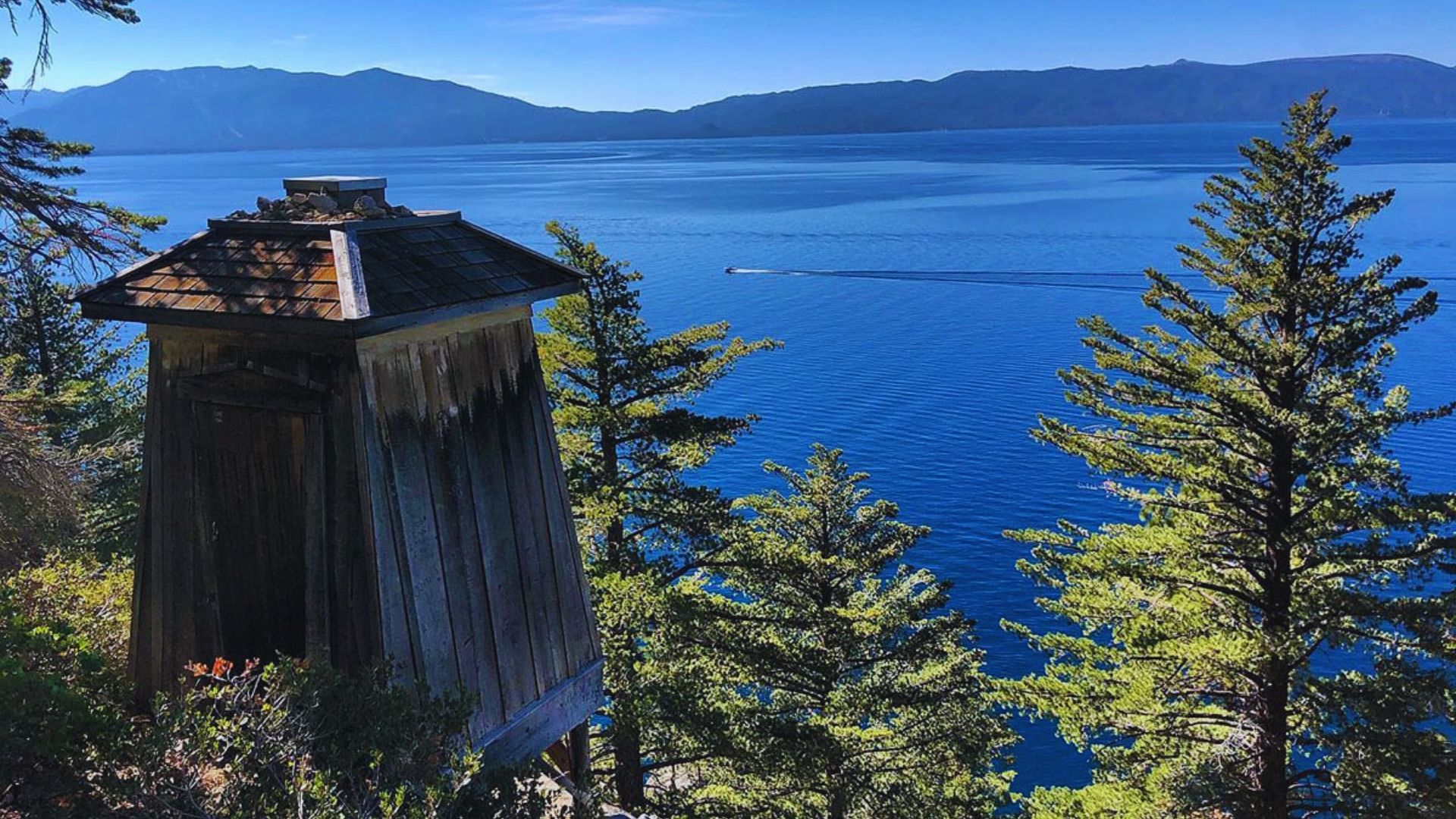 A rustic wooden outhouse with a scenic view of water and mountains in the background.