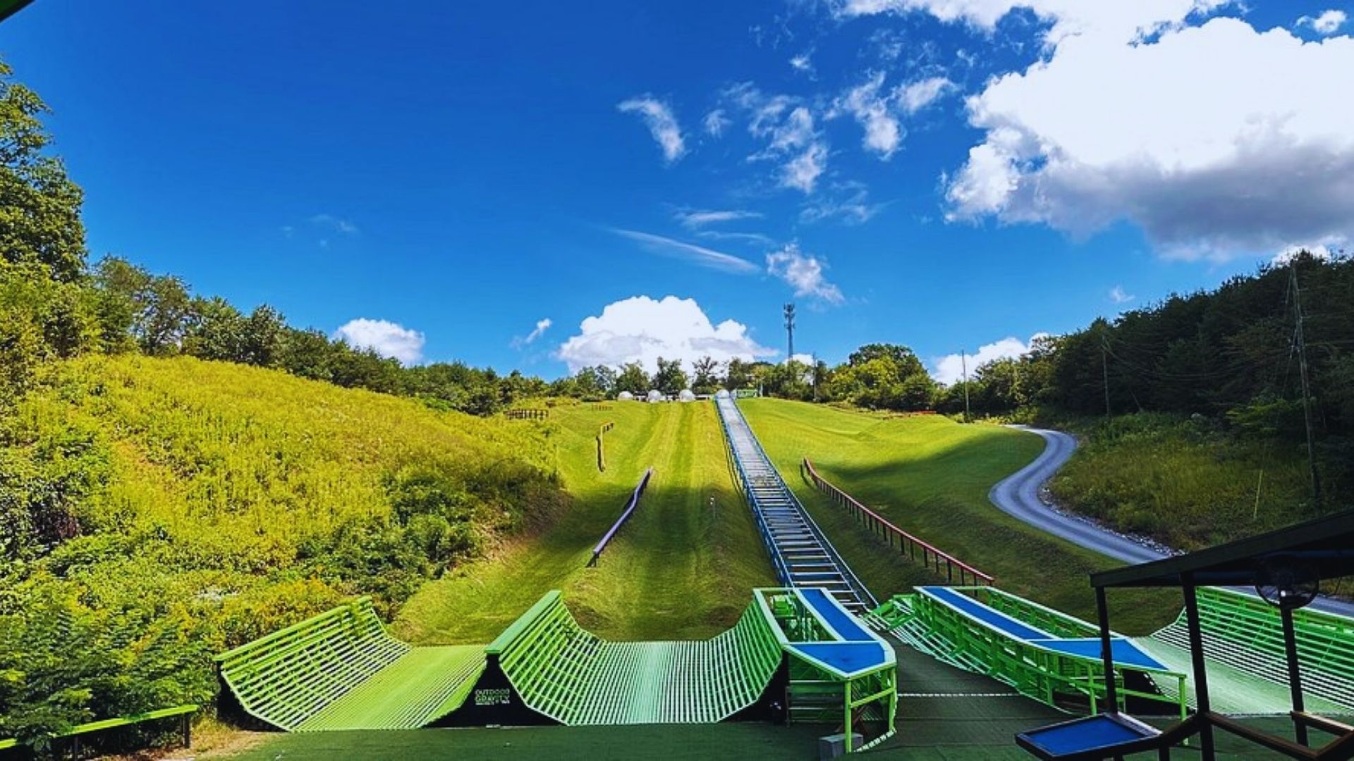 Ski jump hill under a bright blue sky with scattered clouds. Green ramps and tracks lead down the grassy slope surrounded by lush greenery, conveying a sense of readiness and excitement.