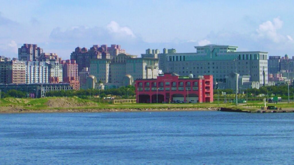 A vibrant cityscape features diverse buildings across a clear blue sky. A distinctive red structure stands by a wide, calm river, conveying urban serenity.