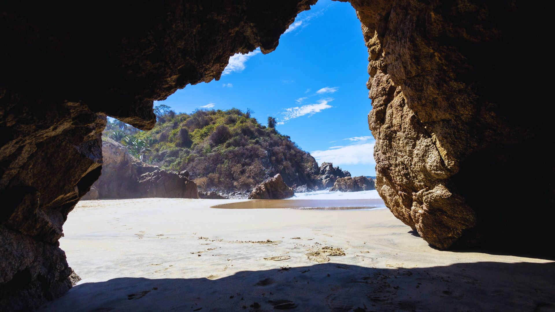 View from a rocky cave opening onto a sunlit beach with clear blue skies. In the distance, waves gently crash on the shore near lush, green cliffs.