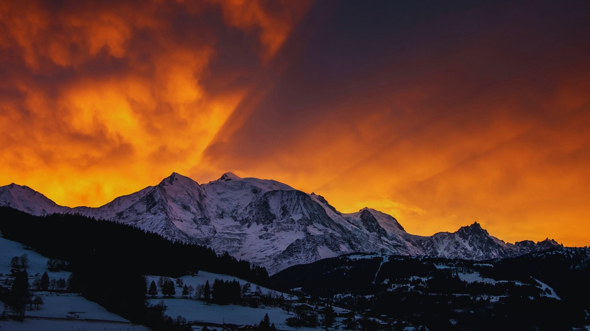 A mountain range illuminated by vibrant orange and red clouds during sunset, creating a dramatic and colorful sky.