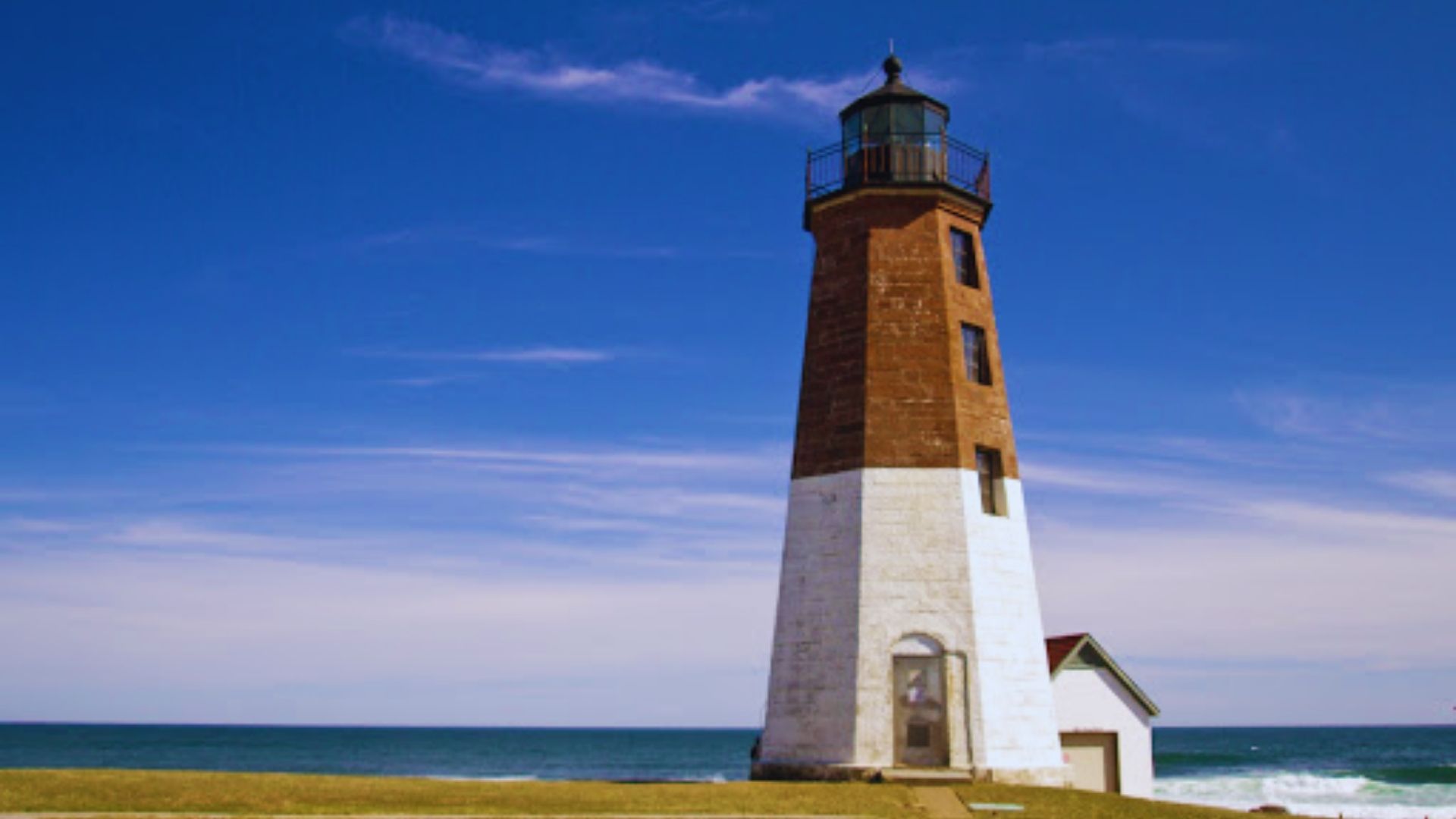 A tall lighthouse stands on a sandy beach, overlooking the ocean under a clear blue sky.