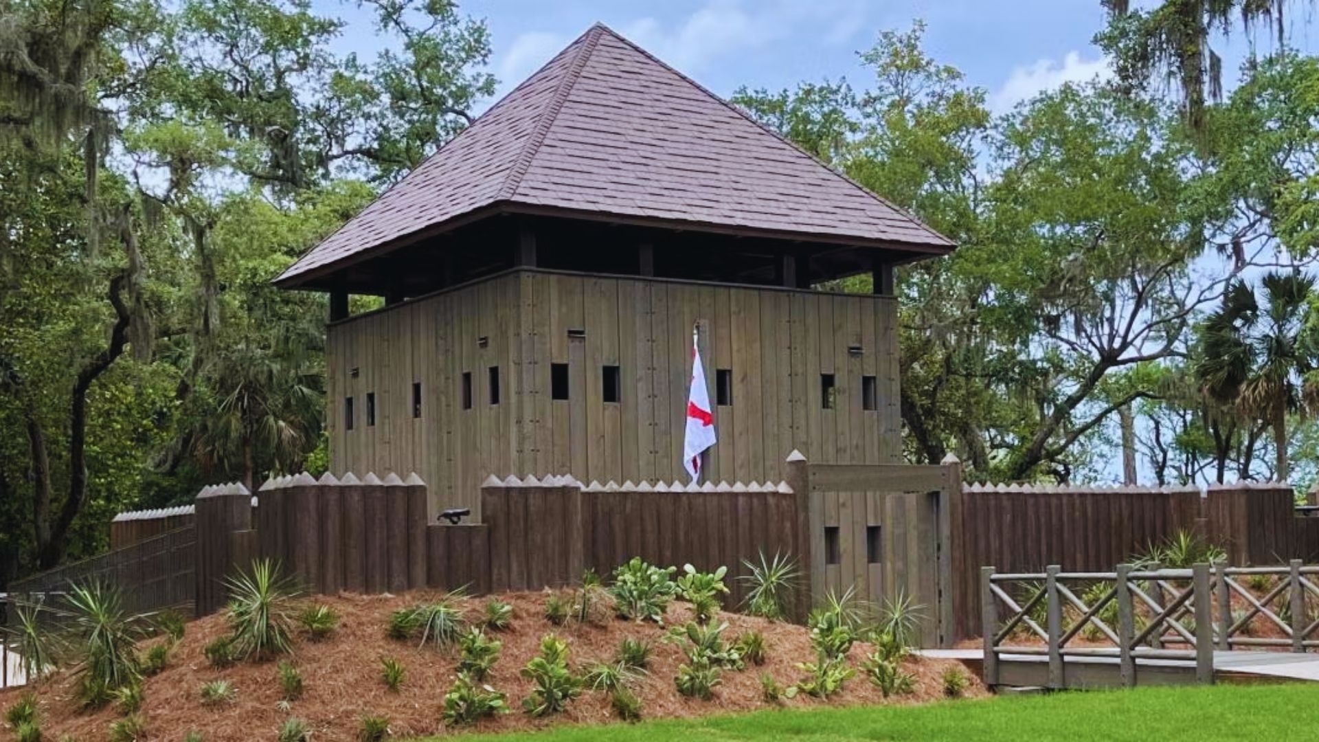 A wooden fort with a tower, surrounded by lush green trees, set against a clear blue sky.