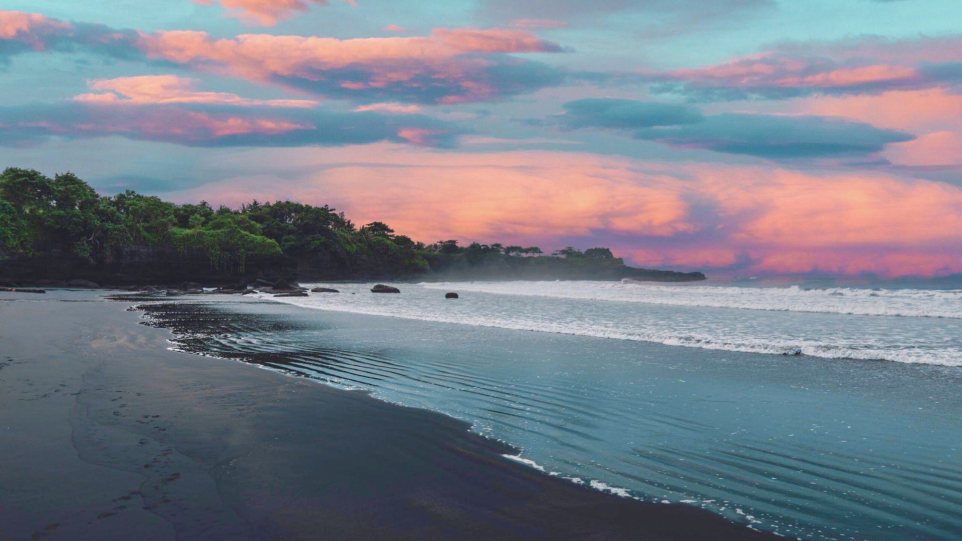 Serene beach with dark sand and gentle waves. Lush greenery lines the shore. Sky painted with pink and orange clouds at sunset, creating a tranquil atmosphere.