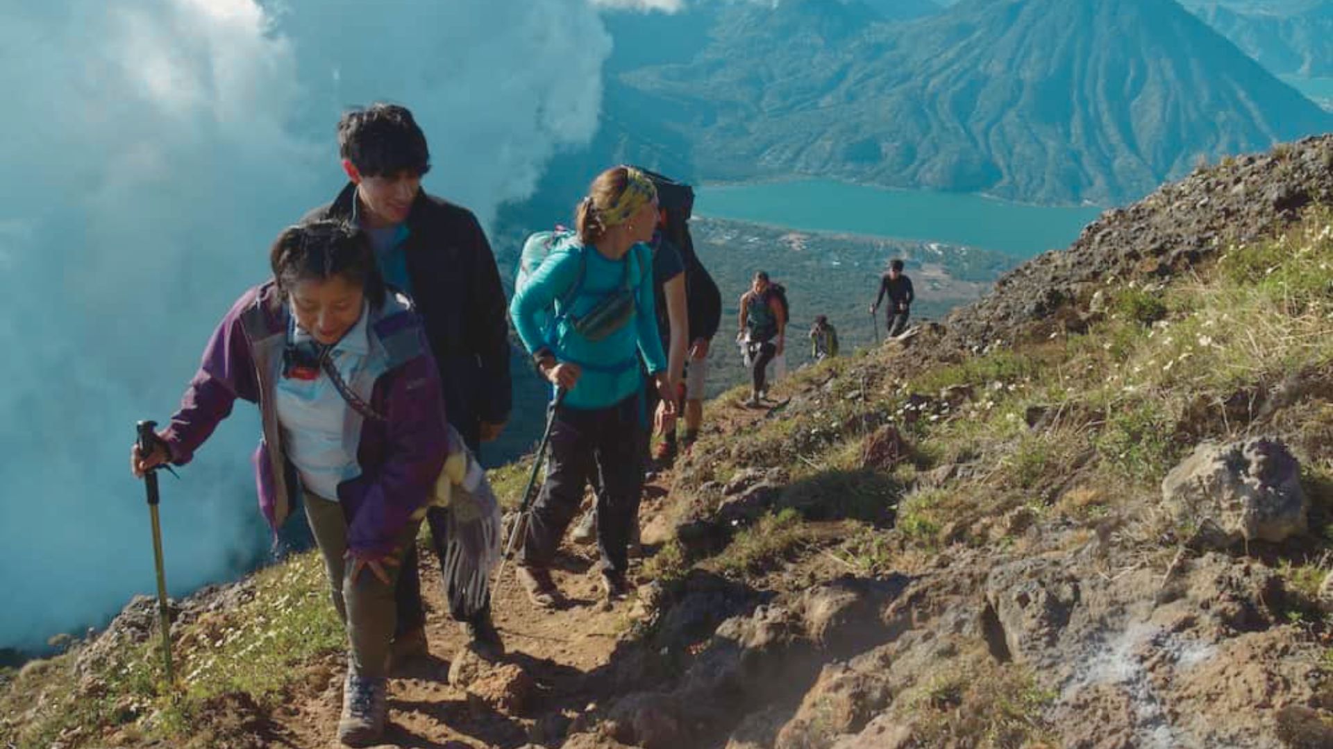 A group of hikers ascends a steep, rocky trail on a mountain, surrounded by clouds. A lake and distant hills are visible, conveying adventure and determination.