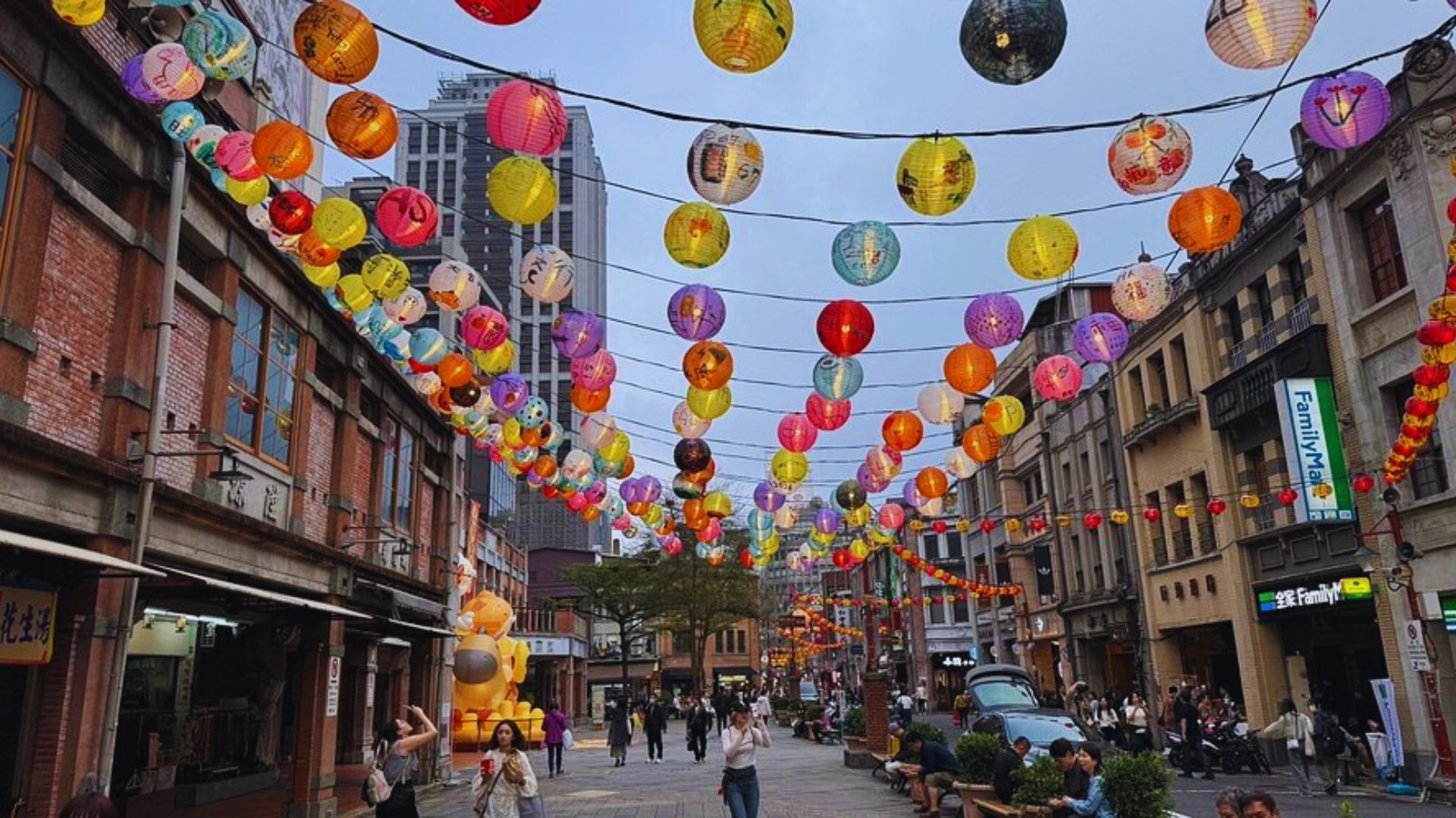 Street scene with colorful paper lanterns hanging overhead, creating a festive atmosphere. People stroll below between historic brick buildings, conveying a lively mood.