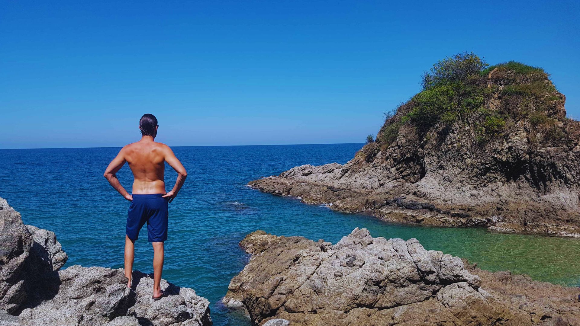A man in swim shorts stands on rocky cliffs, gazing at the vast blue ocean under a clear sky. An island with green foliage is visible nearby.