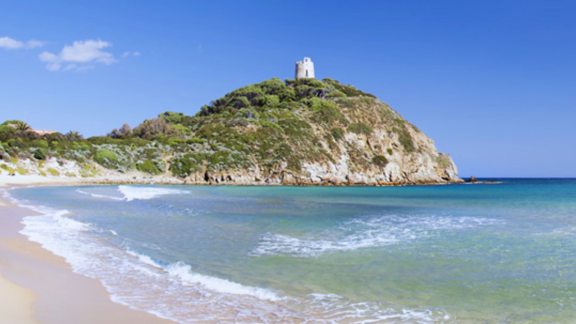 A serene beach with gentle waves and sandy shore is set against a lush, green hill topped by a small white tower. The sky is clear and blue.