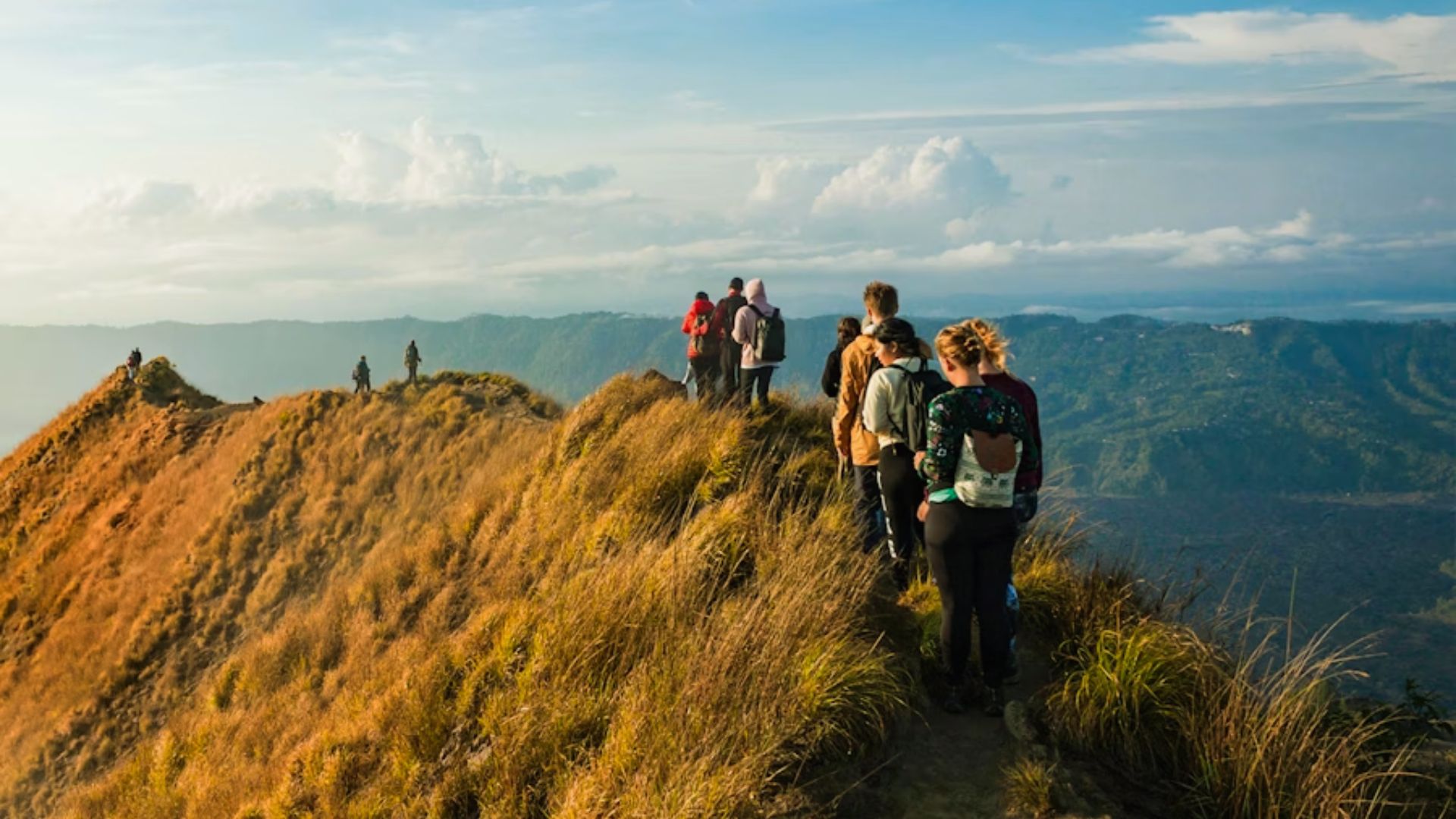 A group of hikers walks along a narrow, grassy ridge on a sunlit mountain with vast views of distant hills and a cloudy blue sky, evoking adventure.