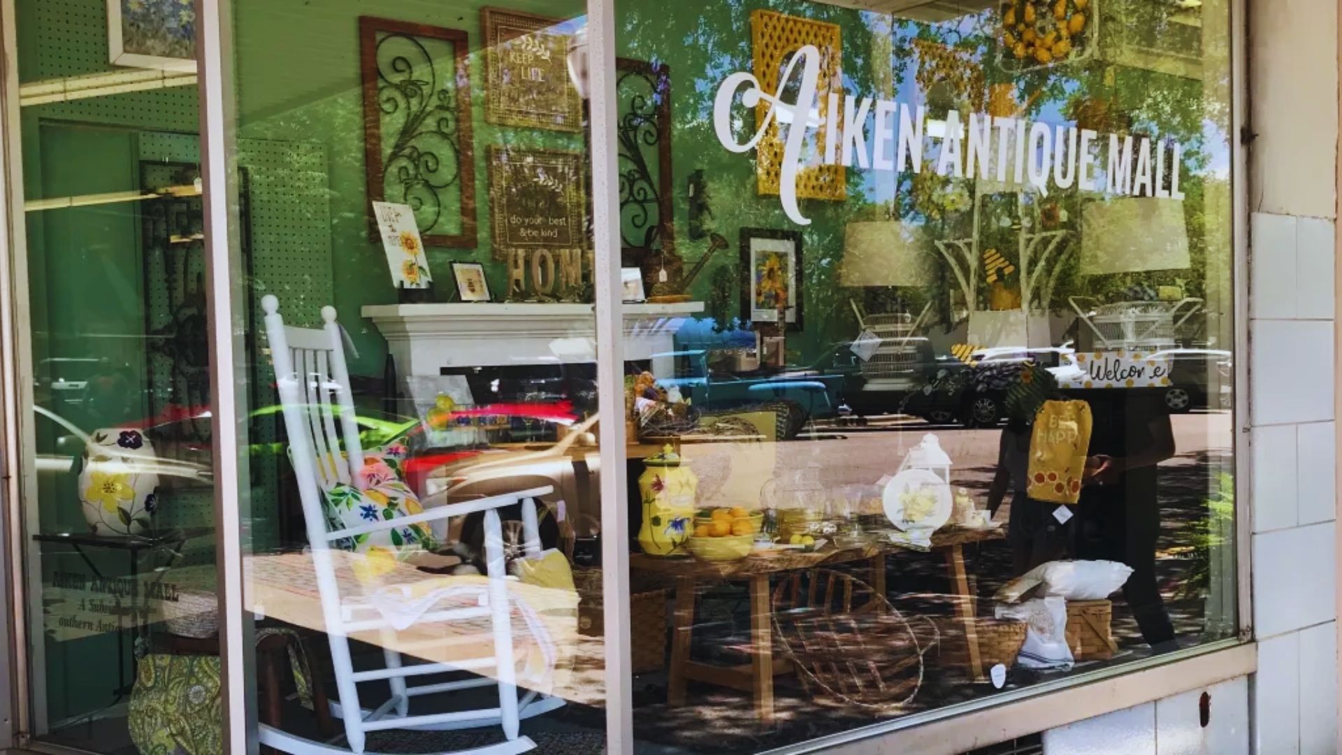 Storefront of Aiken Antique Mall with a reflective glass window displaying vintage decor. Items include a white rocking chair, pillows, and ornamental pieces.