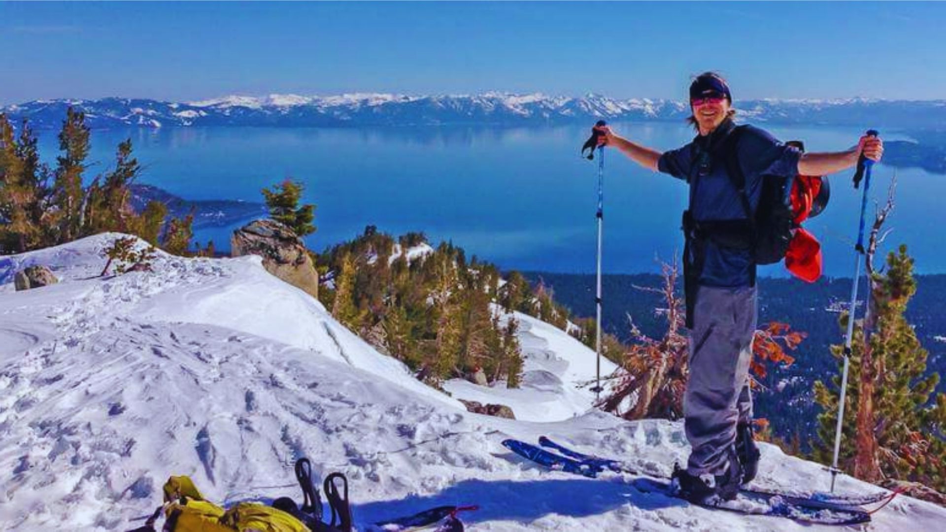 A man on skis stands triumphantly atop a snow-covered mountain, overlooking a vast landscape of peaks and valleys.