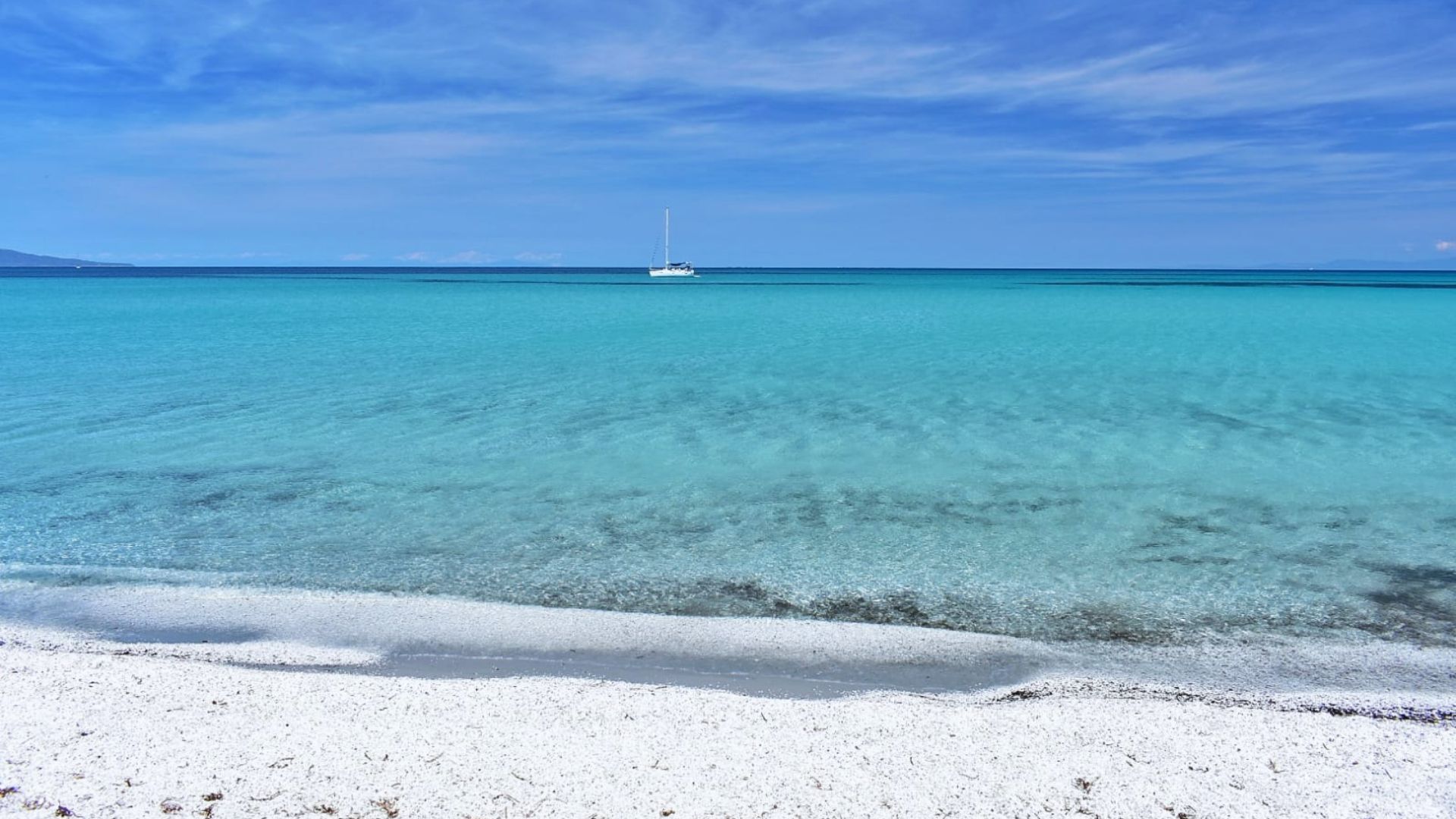 Crystal-clear turquoise sea under a blue sky with wispy clouds, a distant sailboat on the horizon, and a white sandy beach in the foreground. Serene atmosphere.