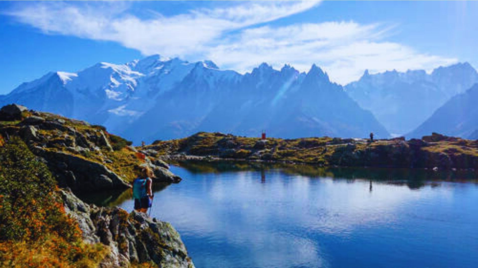 A man stands on a rock, gazing at a serene lake surrounded by majestic mountains in the background.