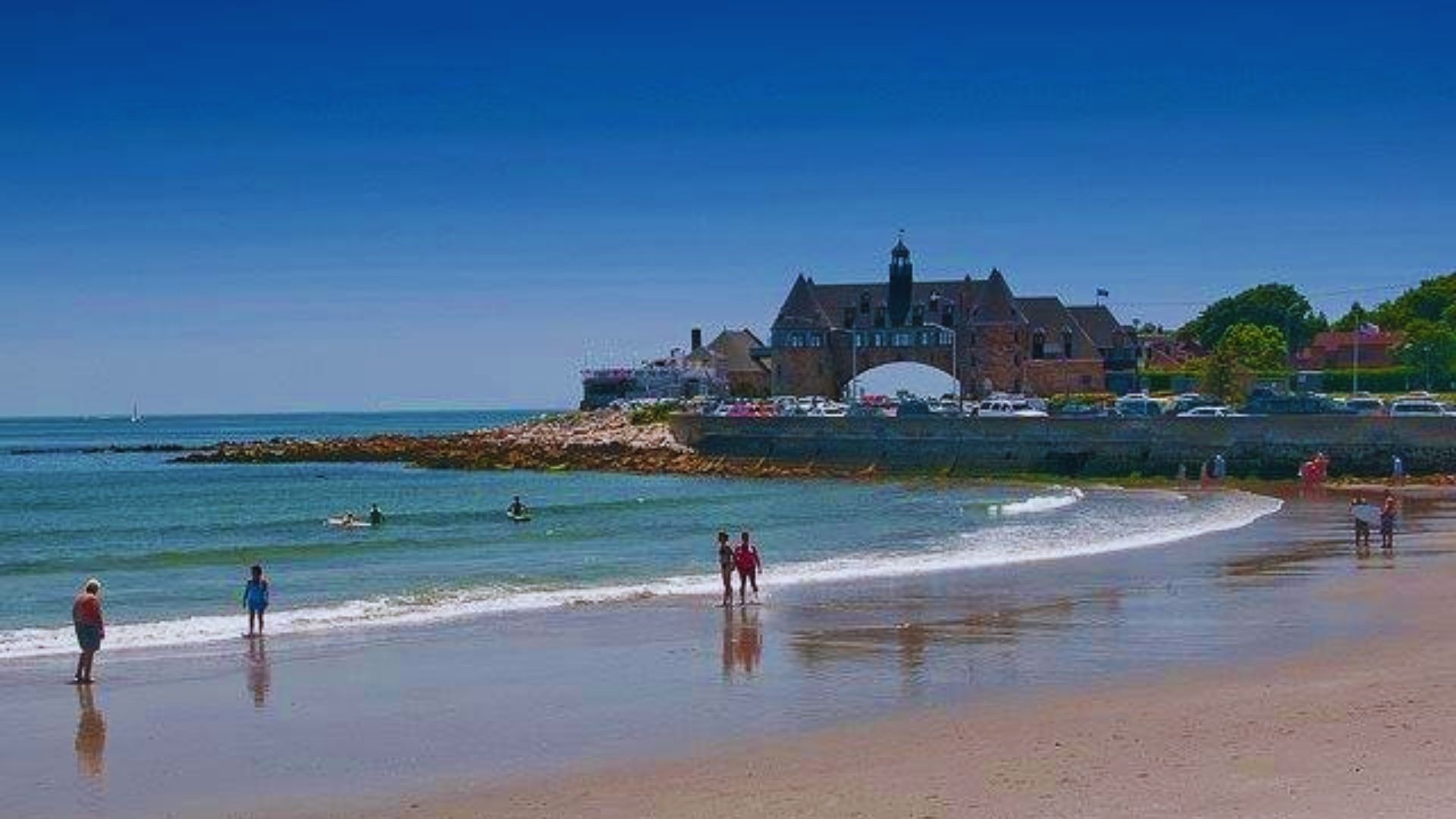 People stroll along a sandy beach, with gentle ocean waves lapping at the shore in the background.
