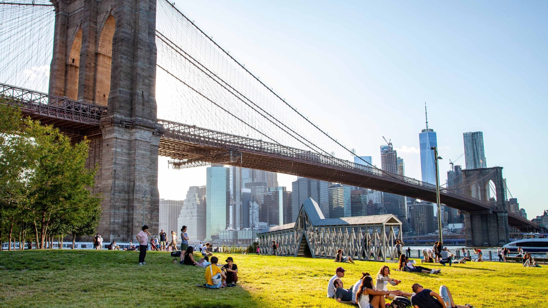 People relax on the grass with the Brooklyn Bridge in the background, enjoying a sunny day in the park.