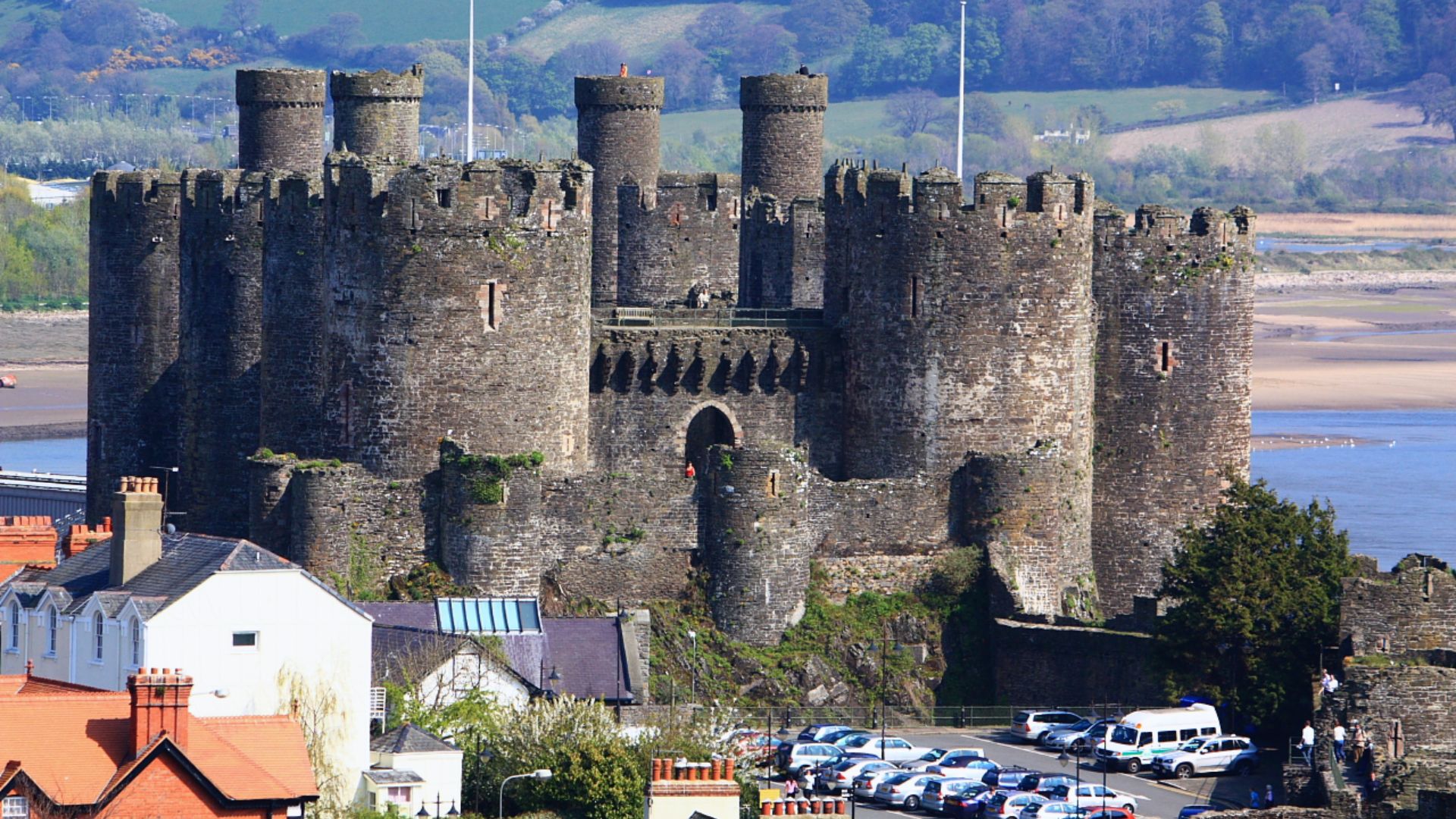 A majestic medieval castle overlooks a river, surrounded by lush hills. Its tall stone battlements exude historical grandeur, with parked cars nearby.