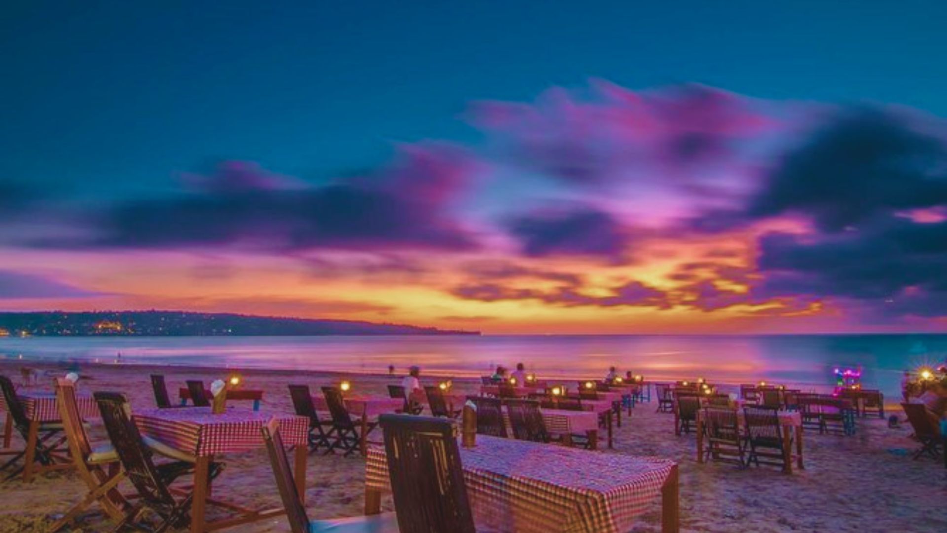 Sunset beach dining scene with tables set on the sand, under a vibrant sky of pink, purple, and orange hues, creating a peaceful and romantic atmosphere.