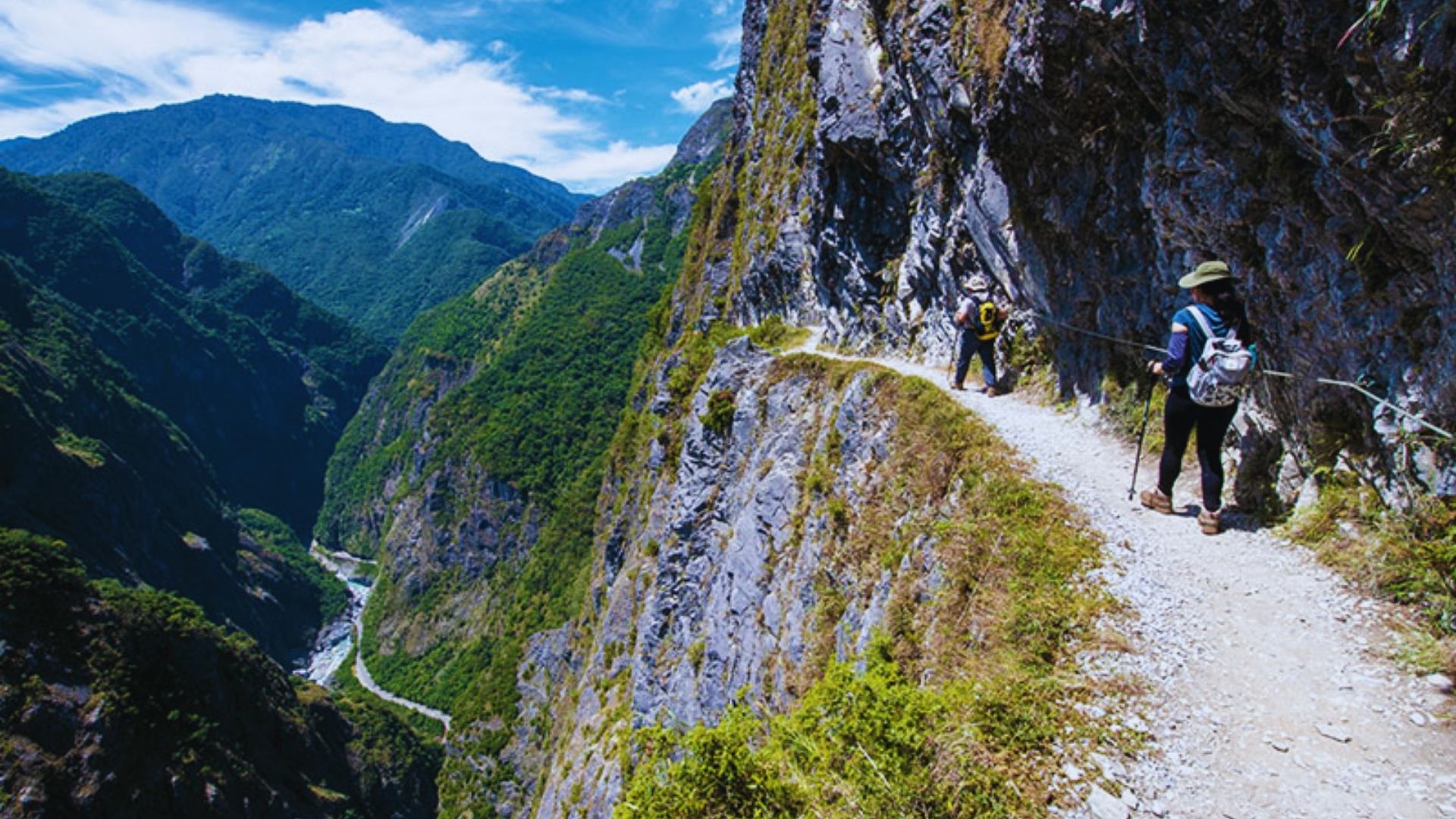 Hikers navigate a narrow, rocky trail on a steep mountain cliff under a clear blue sky. Lush green valleys stretch below, conveying adventure and tranquility.