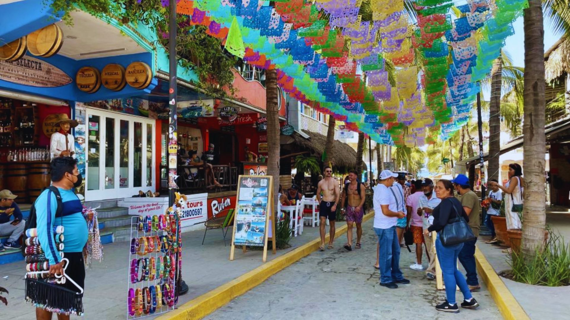 A vibrant street market with colorful papel picado banners overhead. People walk by shops and palm trees, creating a lively, festive atmosphere.